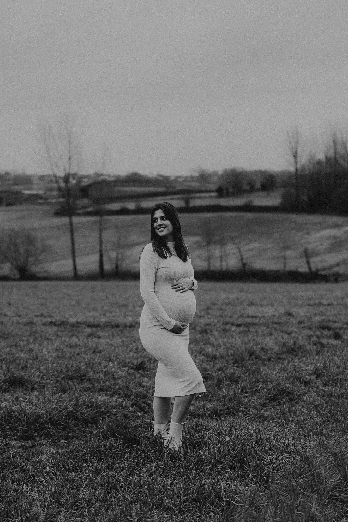 Zwarte en witte foto van een zwangere vrouw die in een open veld staat, met uitgestrekte armen en een glimlach, met een landschap van bomen en heuvels op de achtergrond.