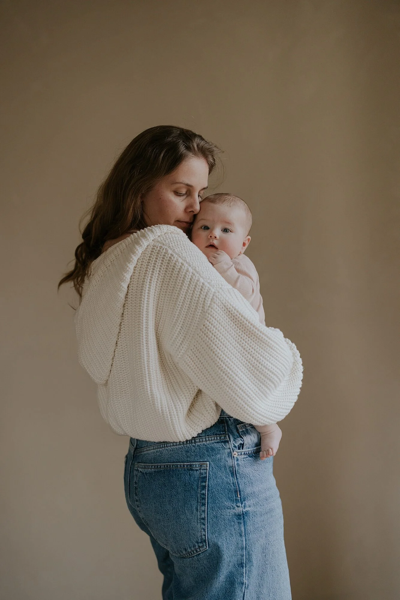 Een vrouw houdt een baby vast, beiden kijken naar elkaar. De vrouw draagt een witte gebreide trui en blauwe jeans, de achtergrond is lichtbruin.