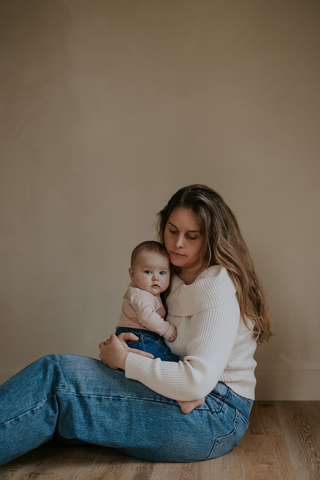 Zittende vrouw met lang haar houdt een baby vast in een lichte kamer met houten vloer, draagt een off-white trui en blauwe jeans.