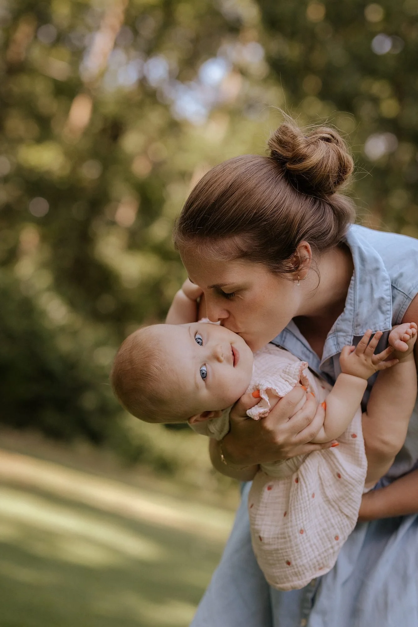 Twee vrouwen, een moeder en een jonge vrouw, knuffelen en kussen een baby met blauwe ogen, buiten in een bosrijke omgeving.