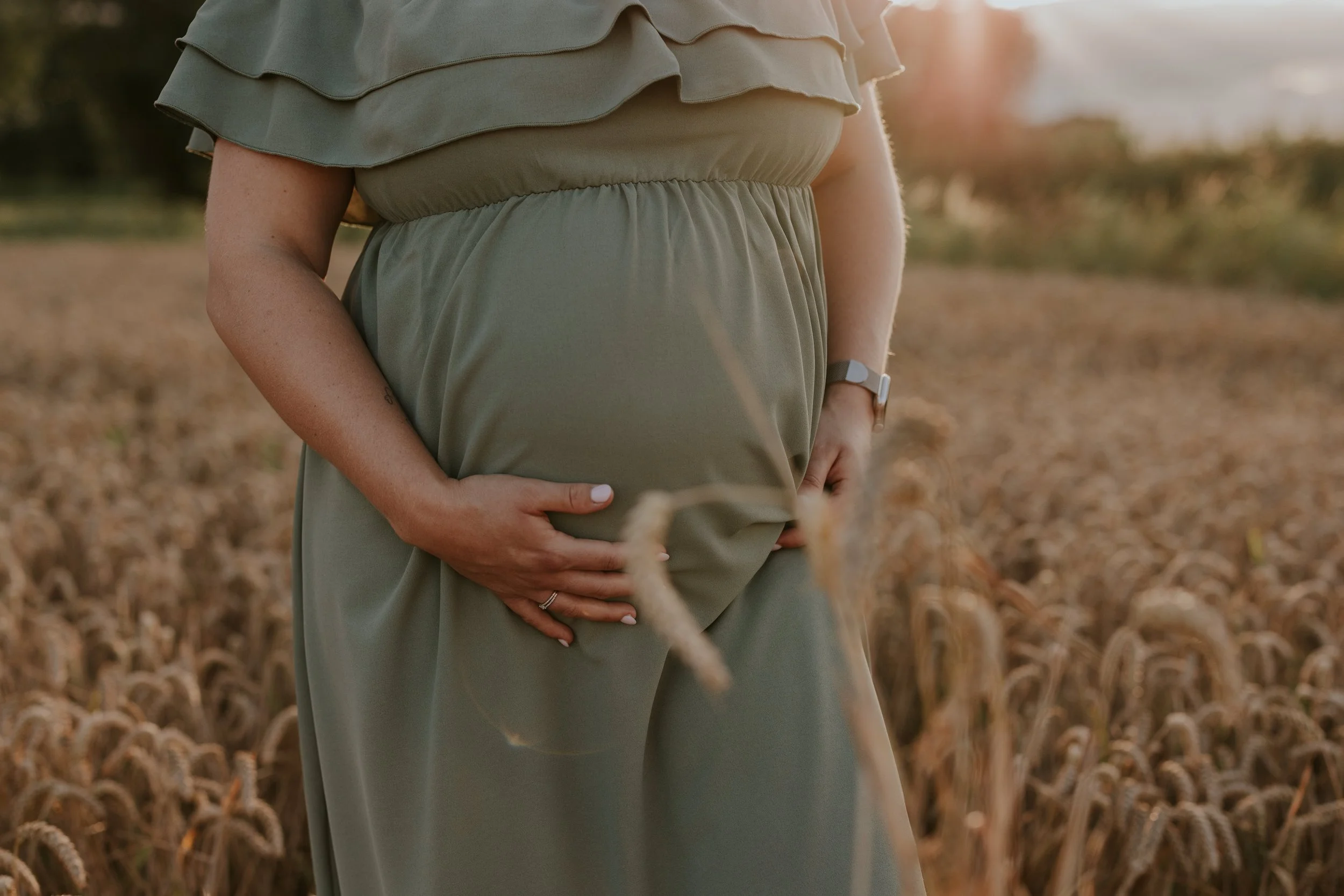 Close-up van een zwangere vrouw in een veld met graan, die haar buik vasthoudt met één hand en haar andere hand op haar buik legt, met zonsondergang op de achtergrond.