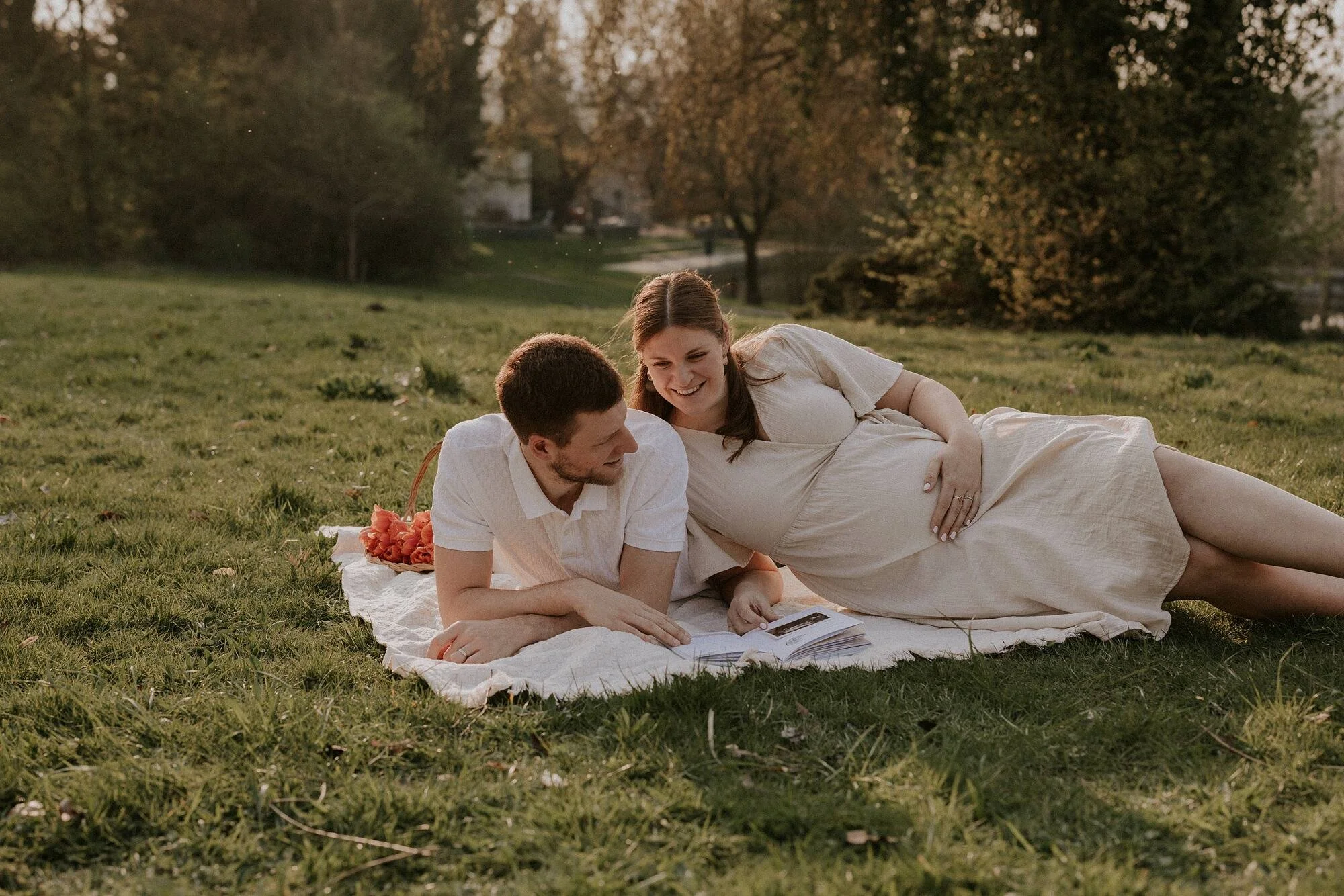 Een zwangere vrouw en een man liggen samen op een grasveld in een park, lachen en lezen een fotoboek.