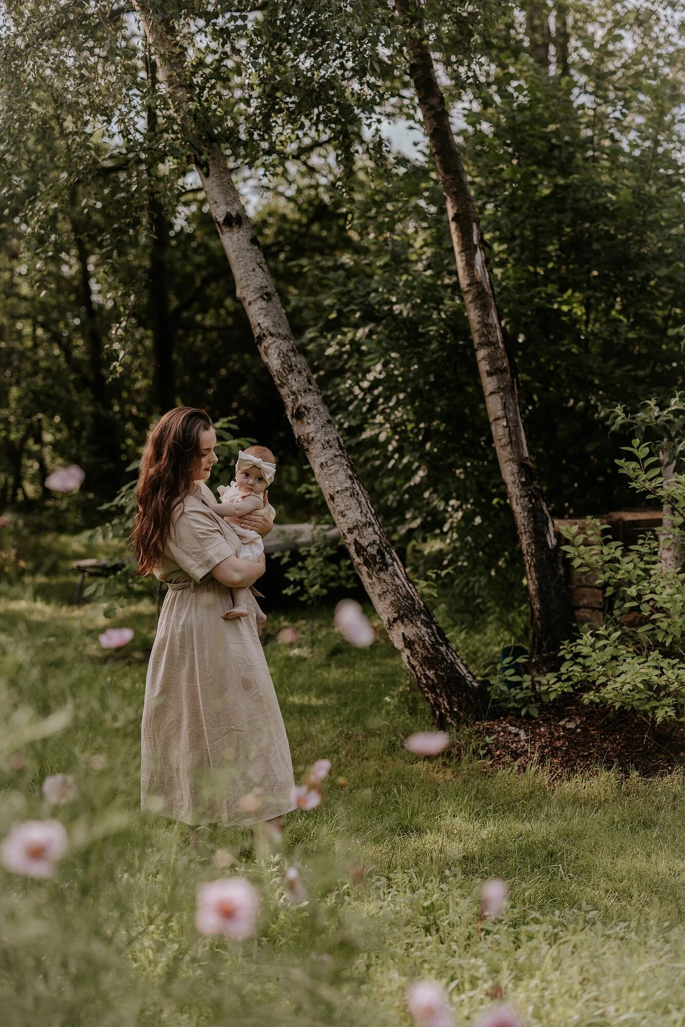 Een vrouw houdt een baby vast in een bosrijke omgeving met groene bomen en grasperken, terwijl ze door bloemen en planten in de foreground lopen.