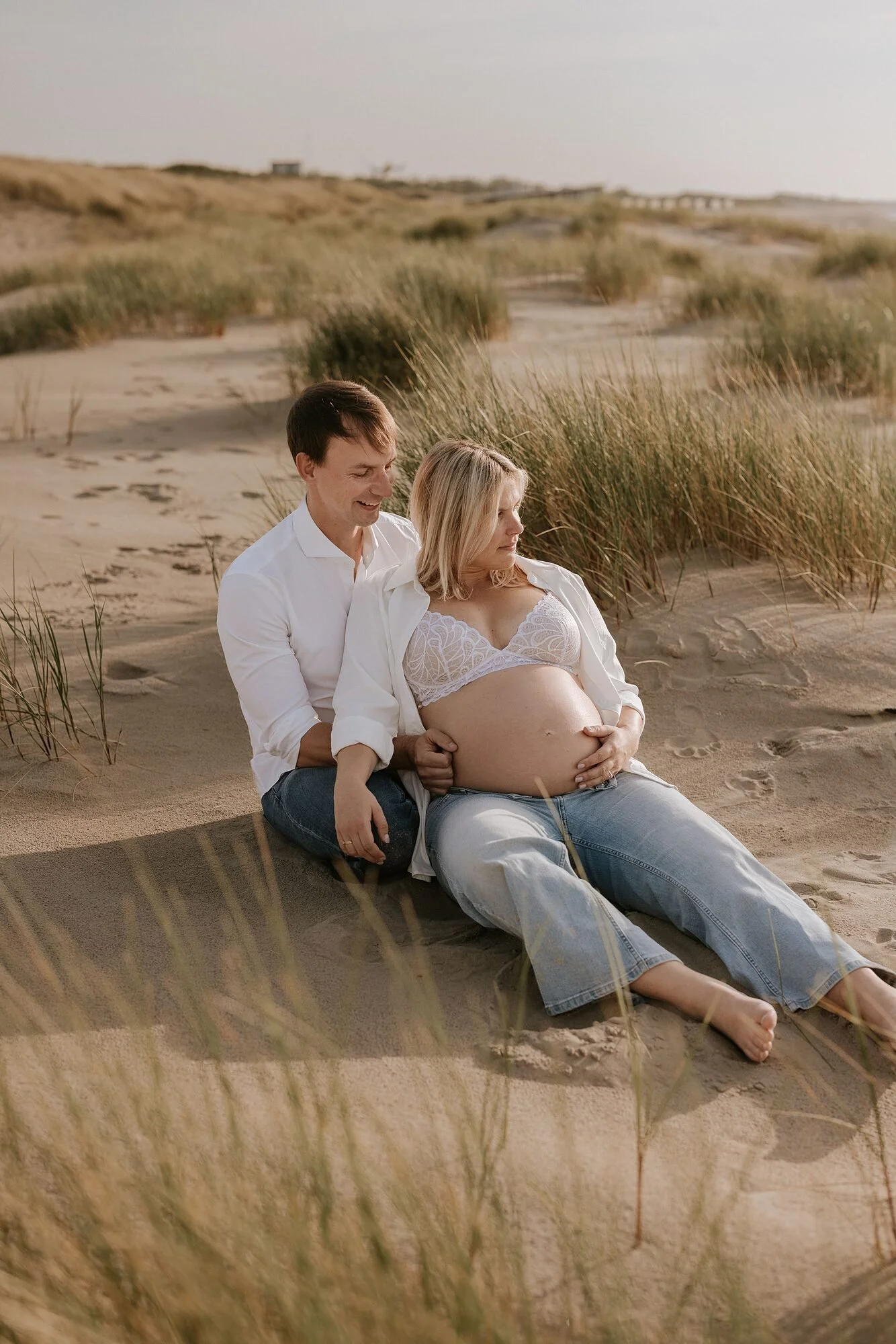 Een zwangere vrouw en haar partner zitten samen op het zandstrand, omringd door gras, met een duinlandschap op de achtergrond tijdens zonsondergang.