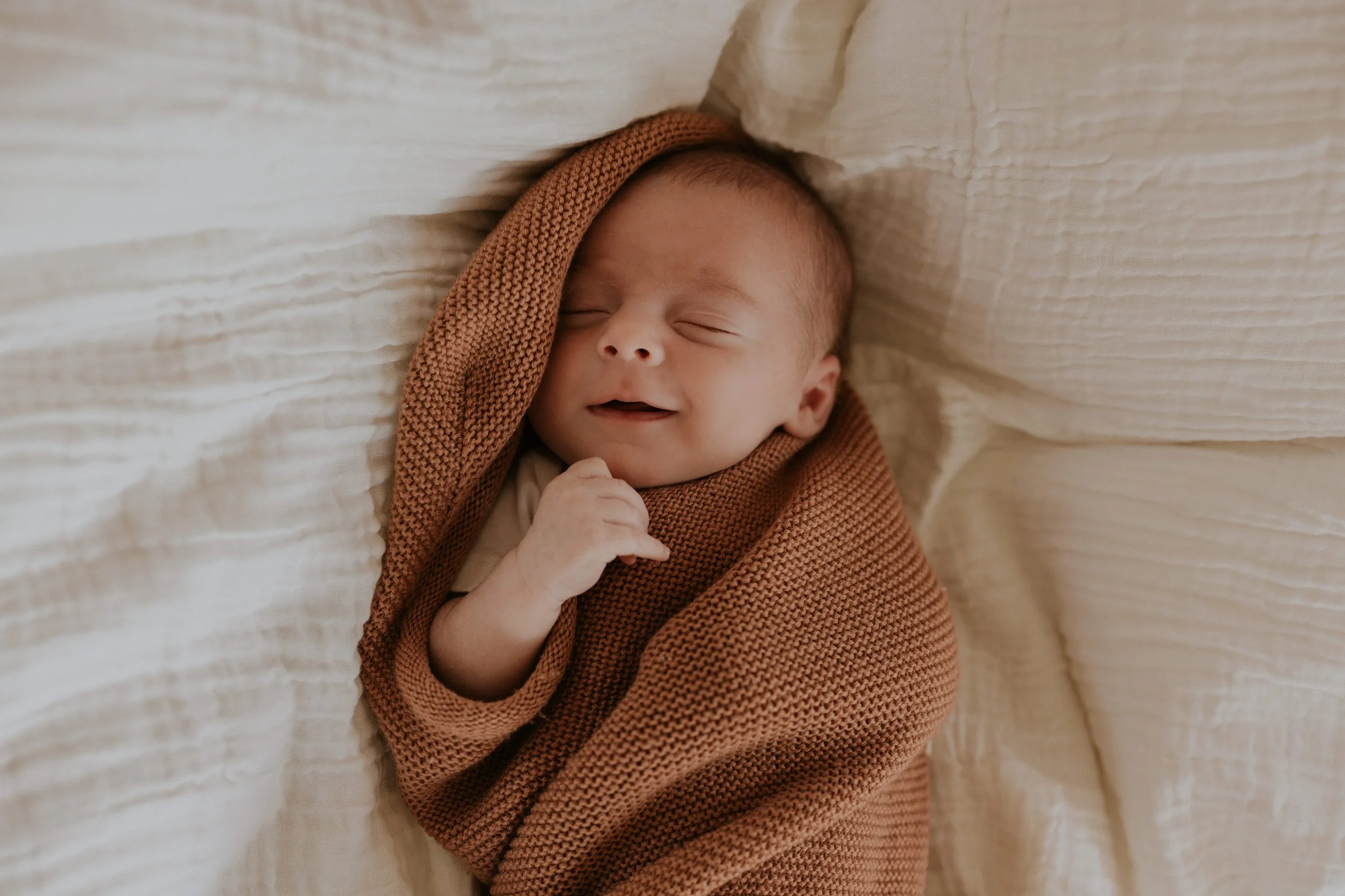 Een baby die ligt in een gewikkeld bruin gehaakt deken, met een glimlach, ogen dicht.