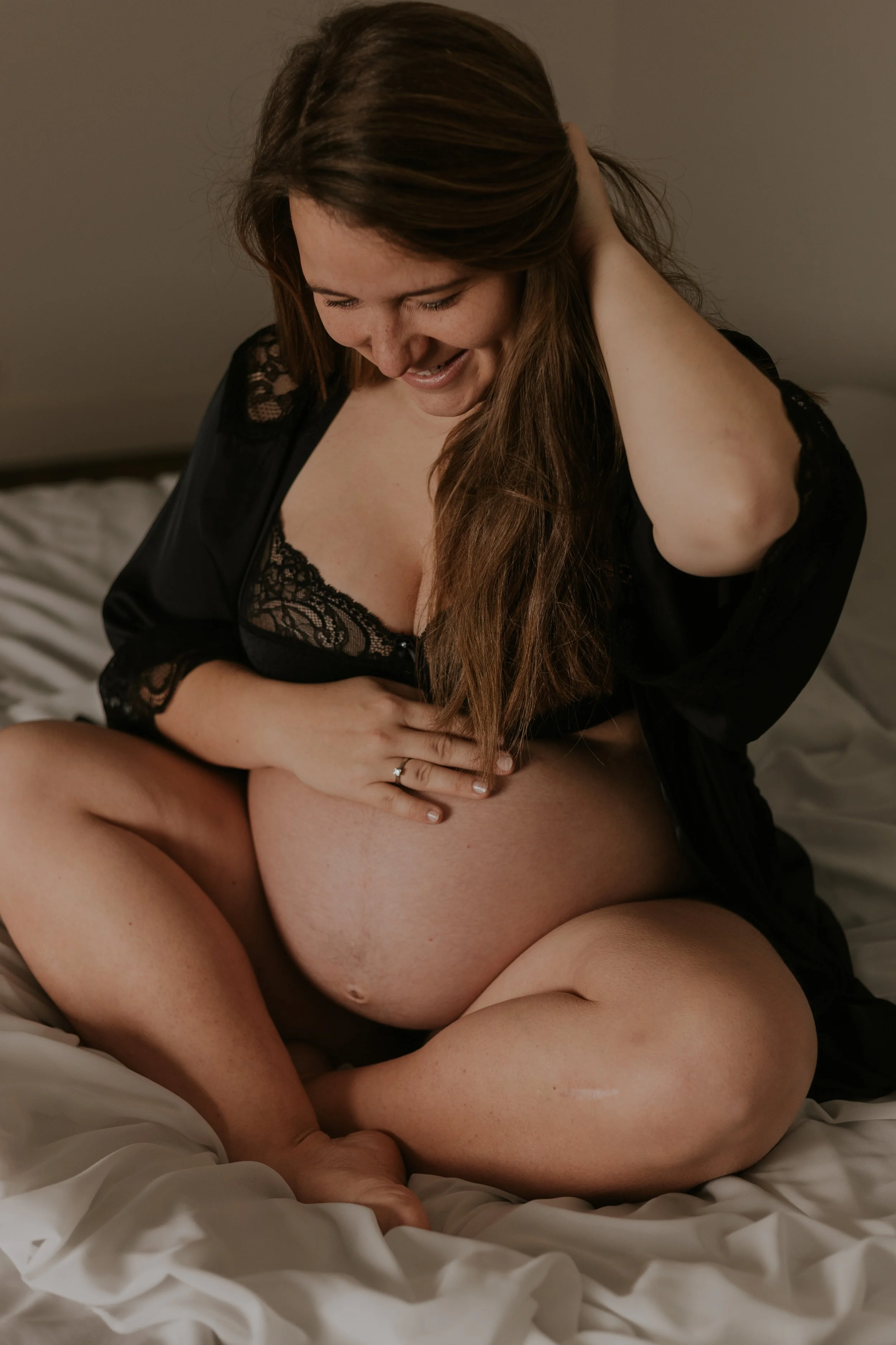 Een zwangere vrouw die op een bed zit, lachend en haar hand op haar buik leggend, gekleed in zwarte kanten nachtkleding.
