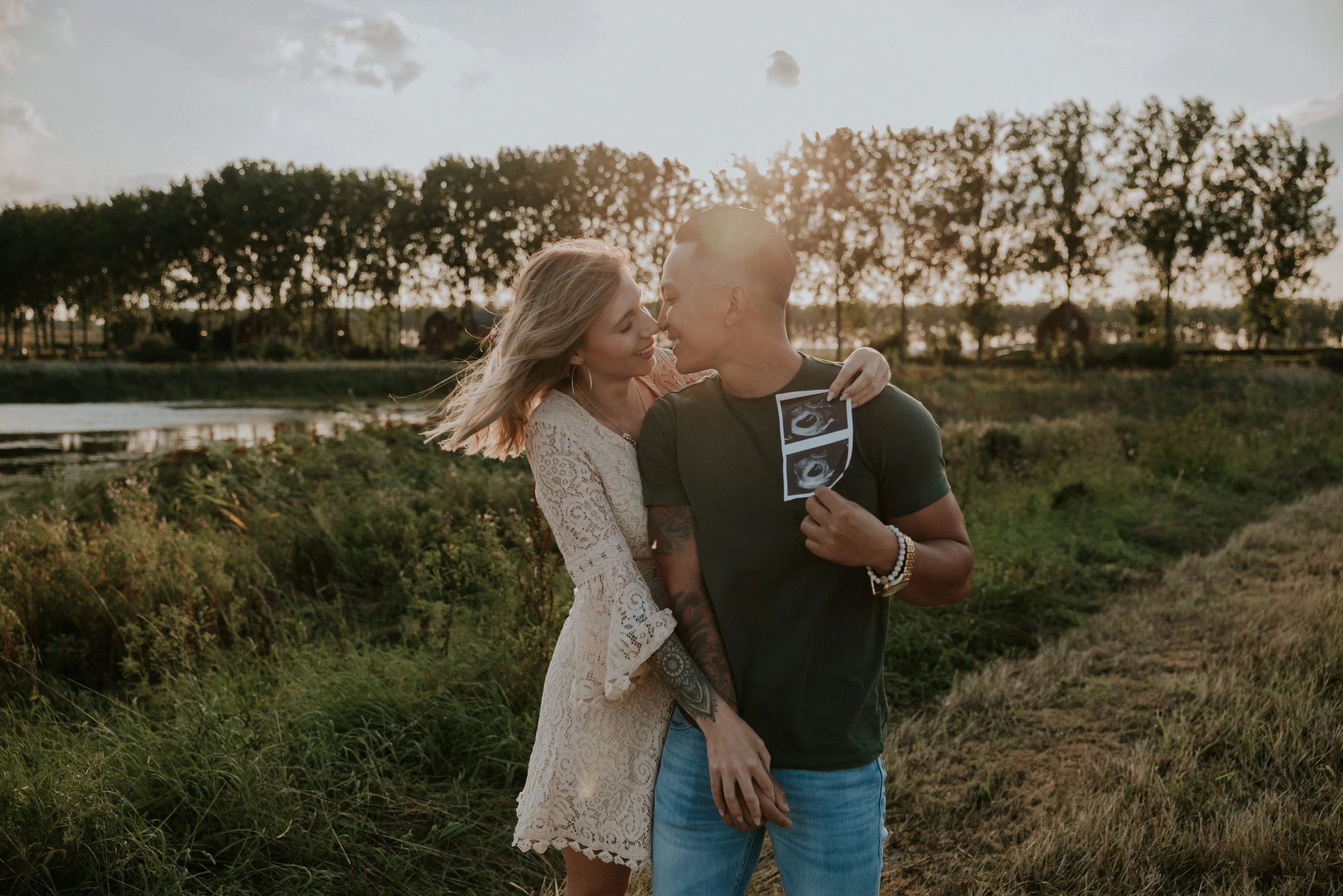 Een koppel dat samen in de natuur staat, met de man die een echo van een early pregnancy toont, en de vrouw die hem knuffelt, beiden glimlachend bij zonsondergang.