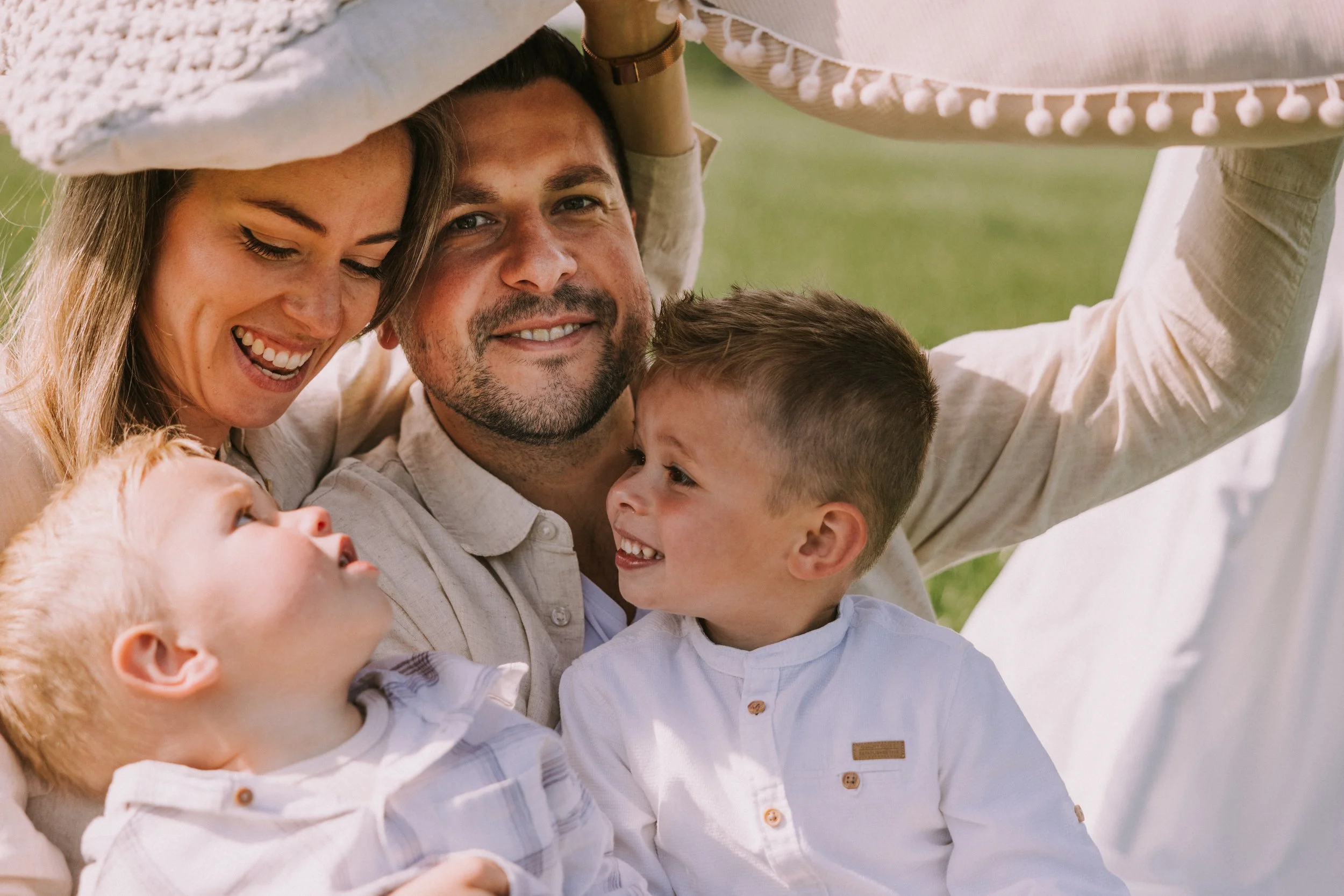 Gezellig gezin dat samen lacht en knuffelt in een groene buitenomgeving, onder een grote parasol.