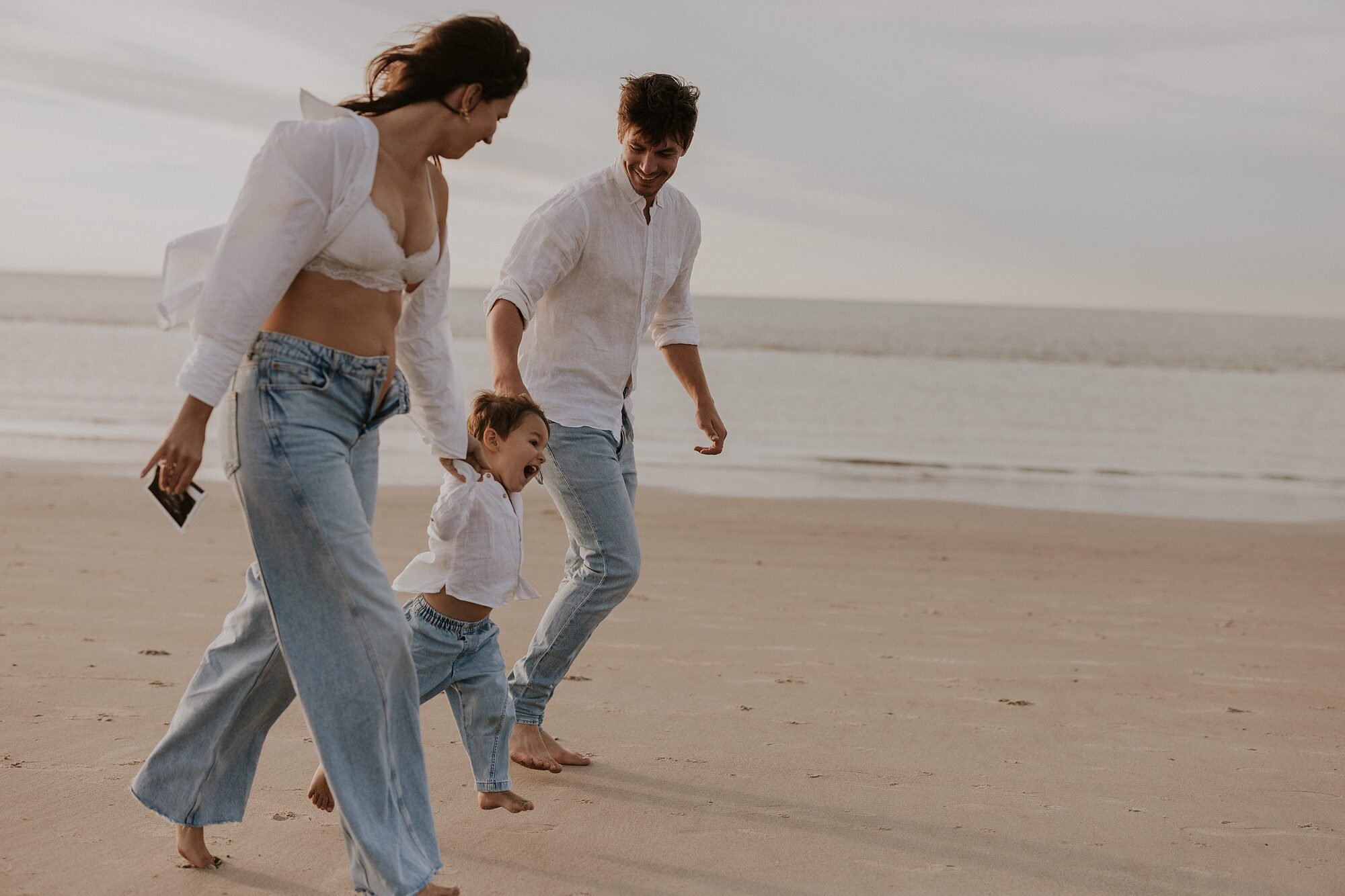 Familie bestaat uit een vrouw, een man en een kind die samen aan het wandelen zijn op het strand. Ze lachen en lijken plezier te hebben terwijl ze dicht bij de waterlijn lopen.