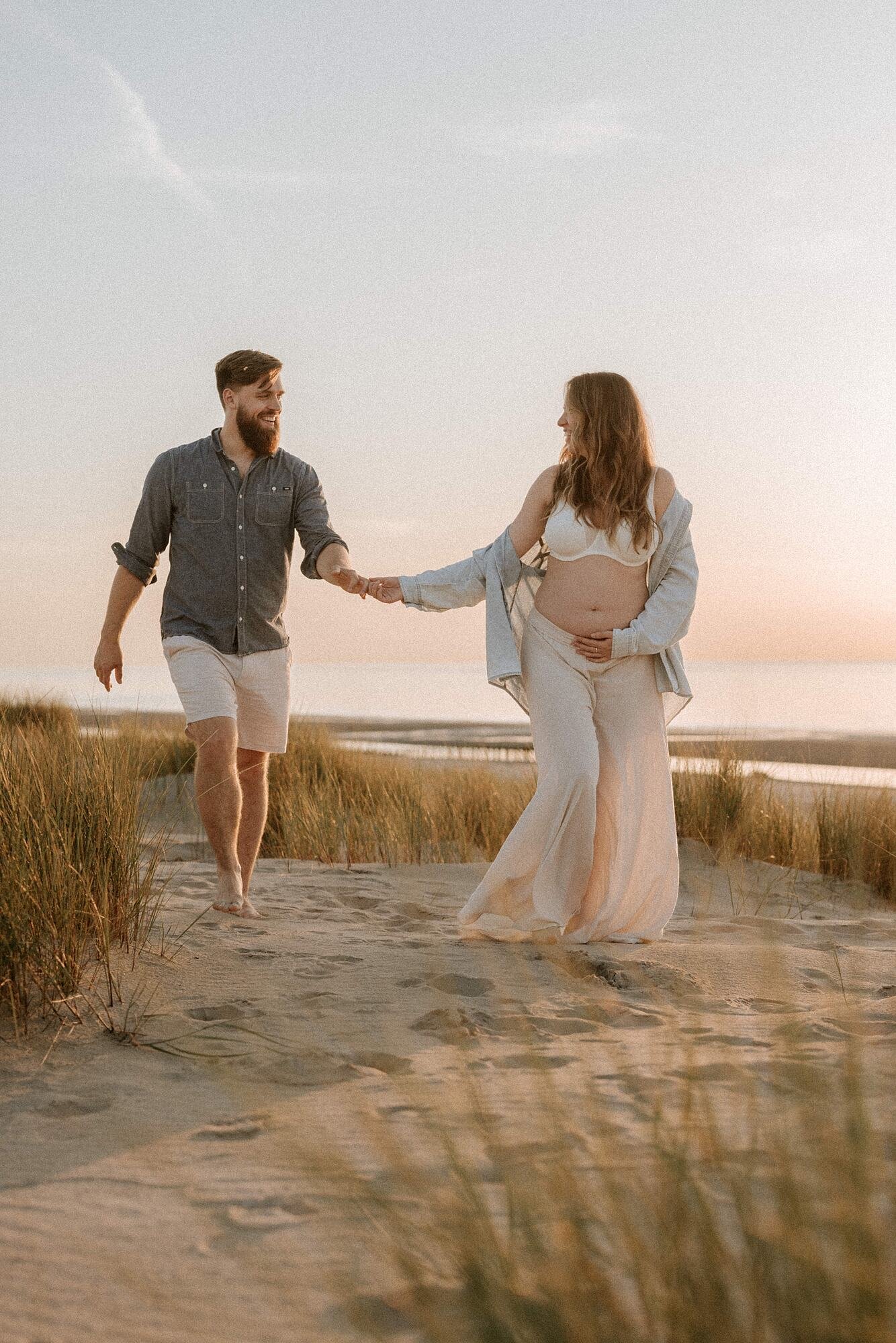 Zwangerschapsshoot aan zee tijdens zonsondergang. Een zwangere vrouw met lang, golvend bruin haar, lachend en met haar hand voorzichtig op haar buik, staat op het strand bij zonsondergang.