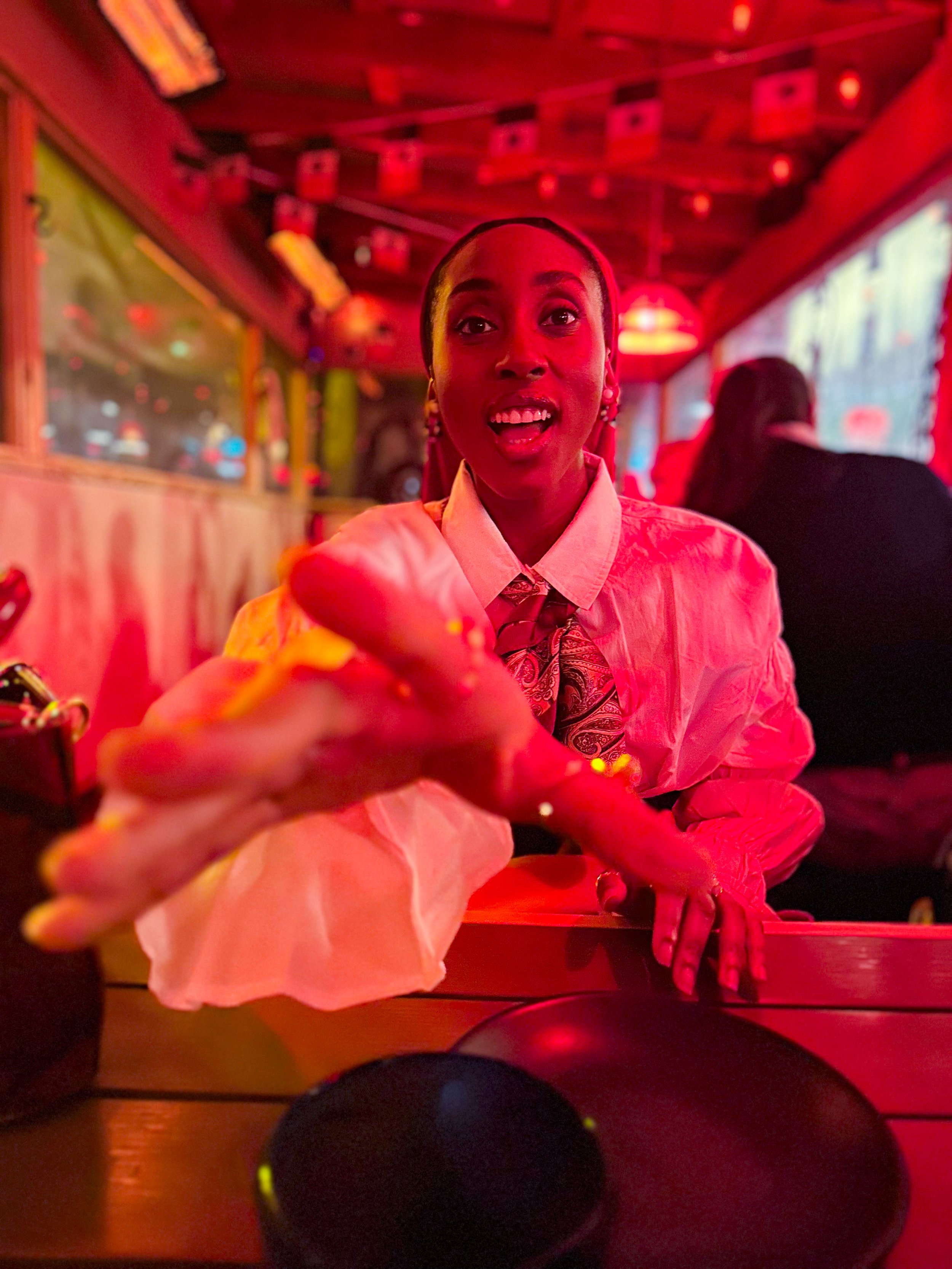 Adaeze in a white shirt and patterned tie reaches out towards the camera with a surprised expression inside a restaurant with red lighting.