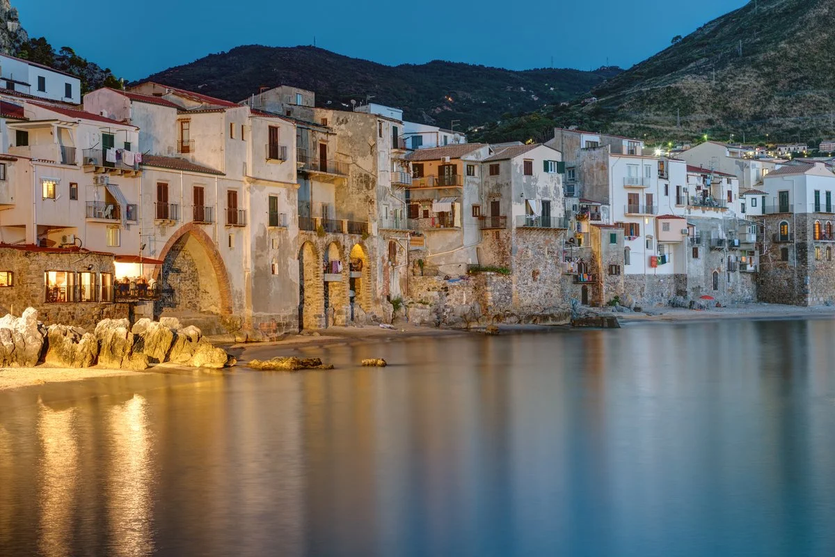 Vista di una spiaggia attraverso un arco in pietra di un edificio storico, con un molo che si estende in acqua e persone che camminano o si rilassano sulla spiaggia.
