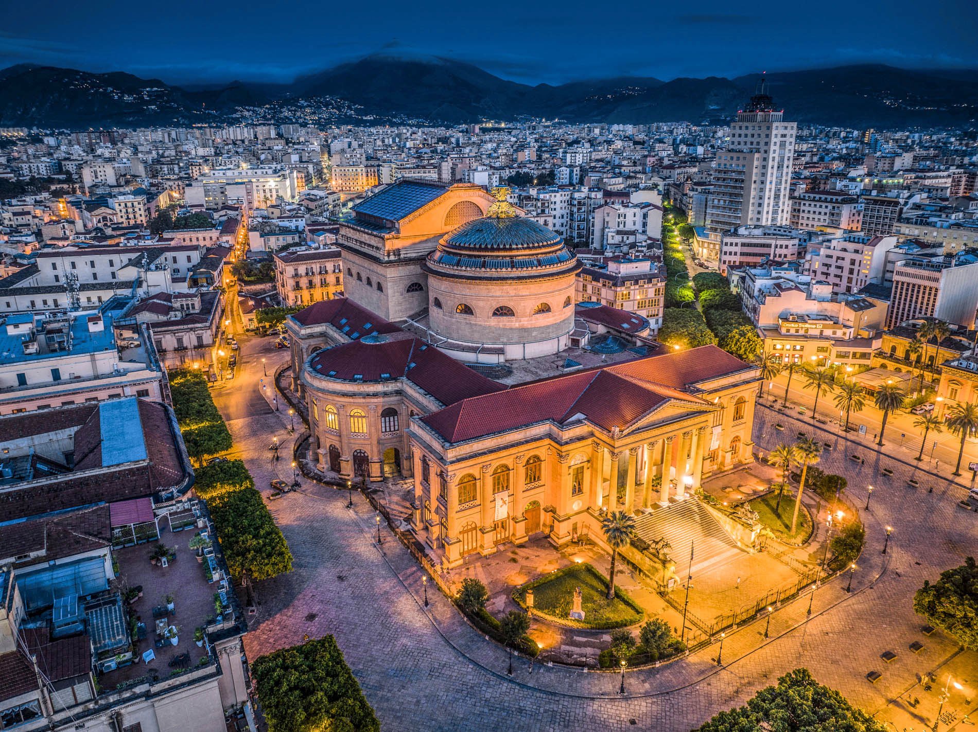 Veduta aerea di Napoli al tramonto con il Teatro di San Carlo illuminato, al centro, e il panorama dei Monti Lattari sullo sfondo.