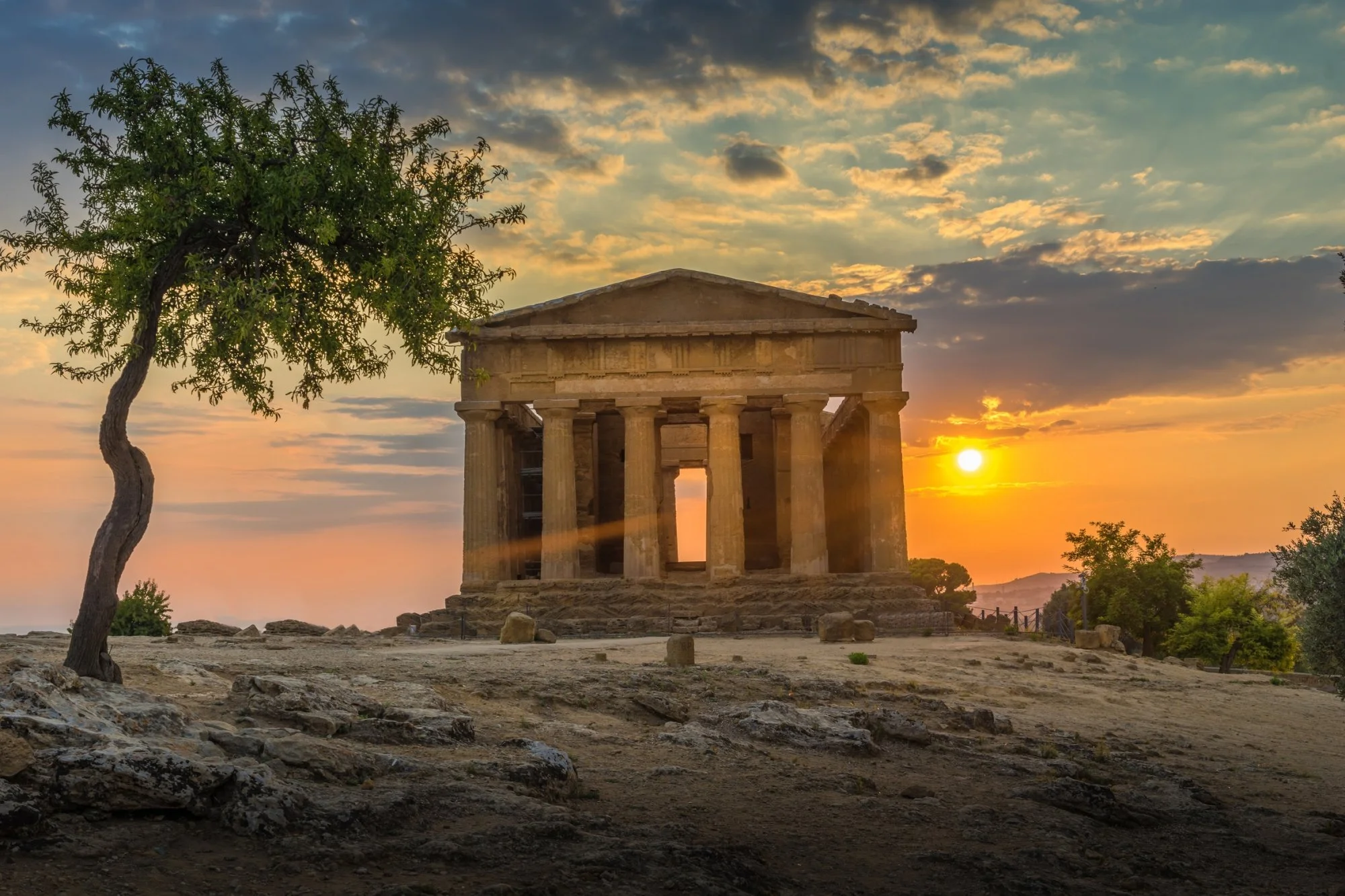 Un antico monumento in stile greco con colonne, al tramonto, con un albero e un cielo colorato sullo sfondo.