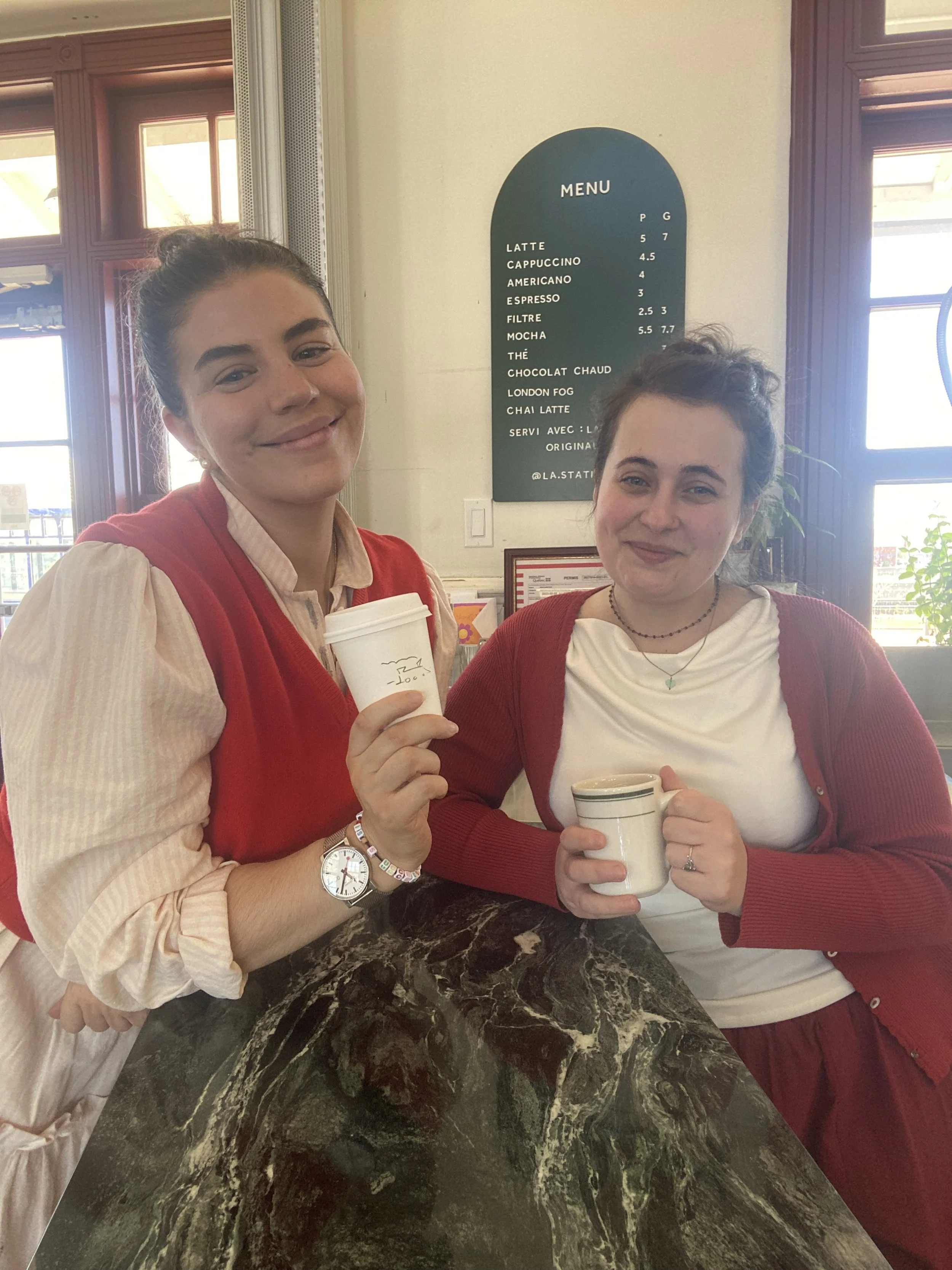 Two female business-owners standing inside their vegan coffee shop, holding cups of coffee and smiling at the camera.