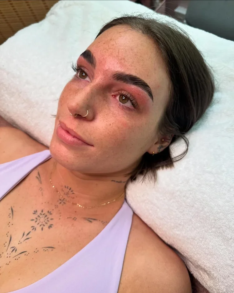 A woman lies on a white towel, wearing a lavender top. Her eyebrows appear freshly groomed, and she has subtle tattoos on her chest, exuding a calm and serene vibe.