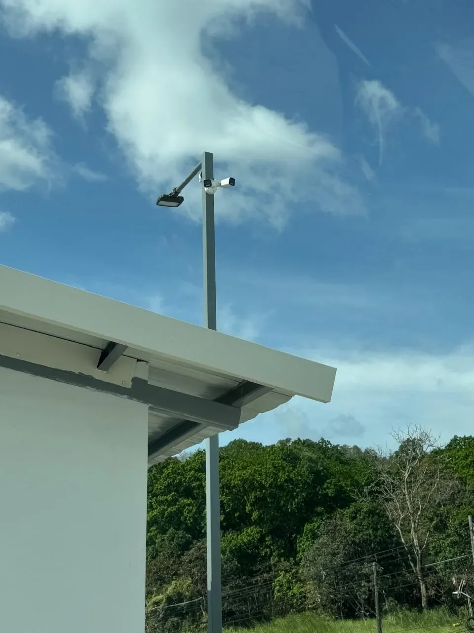 Callejón con poste de iluminación y cámara de vigilancia, cielo con nubes y árboles verdes en el fondo.