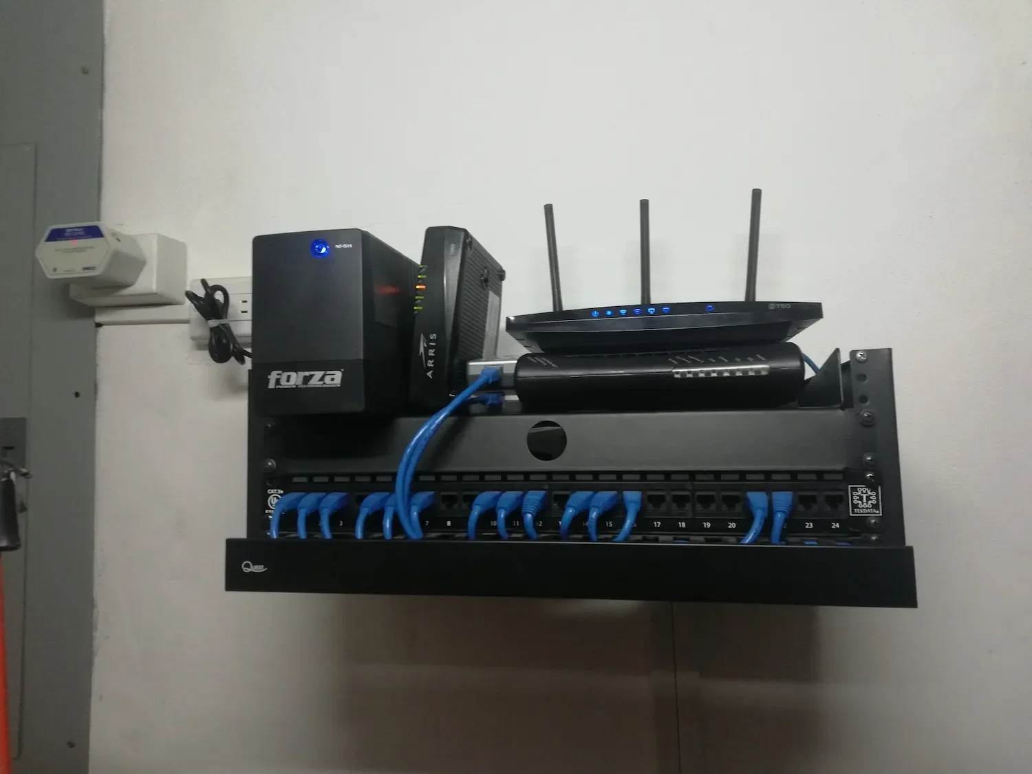 Panel de conexiones con cables azules, un router con antenas, un punto de acceso y un tipo de inversor o servidor en una pared blanca.