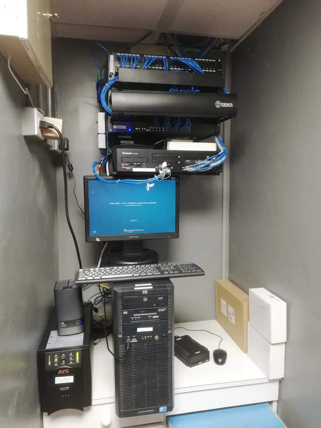 Equipo de tecnología y redes con un monitor, teclado, CPU, y componentes de cableado en un gabinete de servidores.