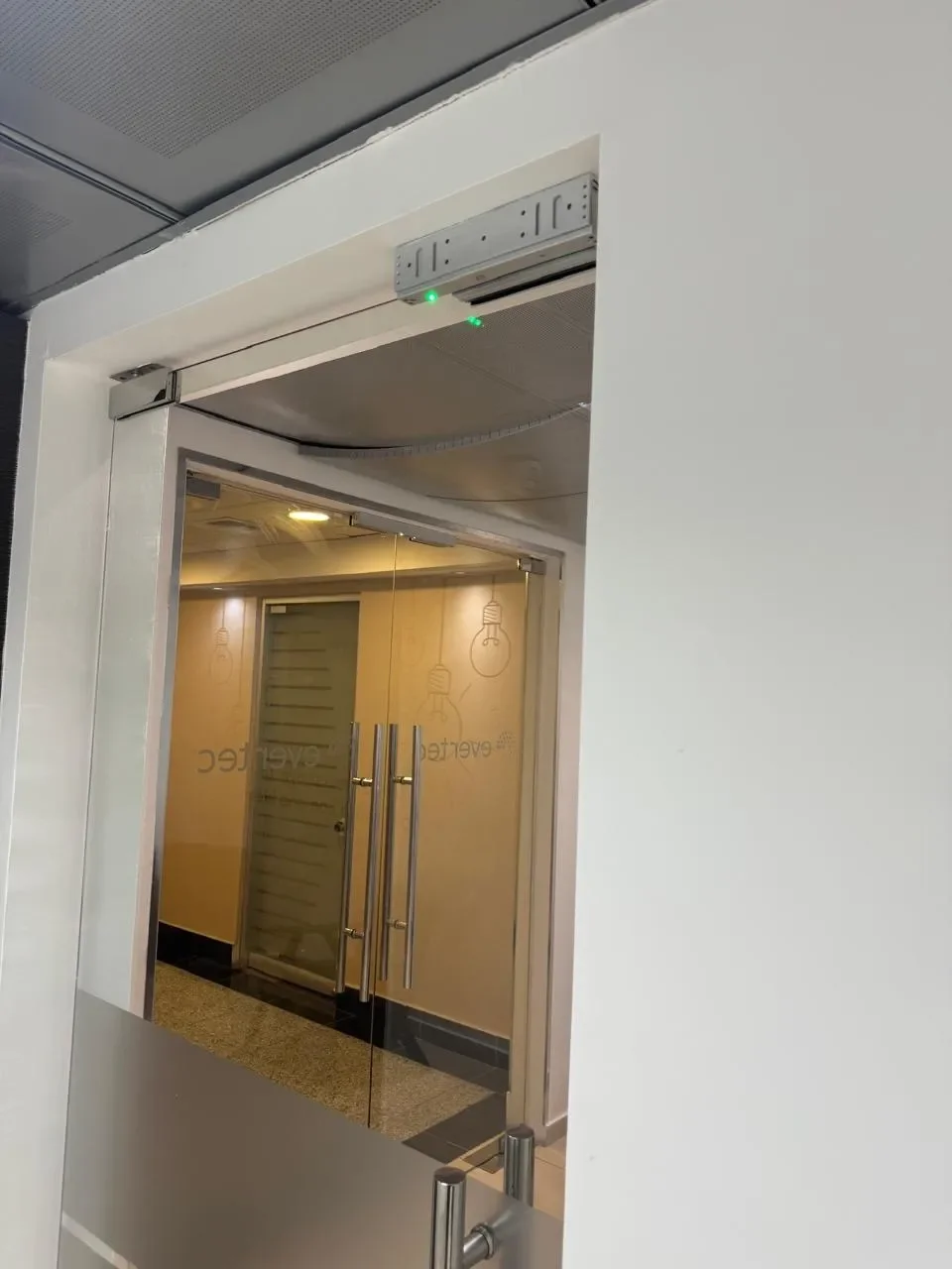 Puerta de acceso a una oficina con doble mampara de vidrio y un sistema de seguridad en la parte superior, dentro de un edificio moderno.