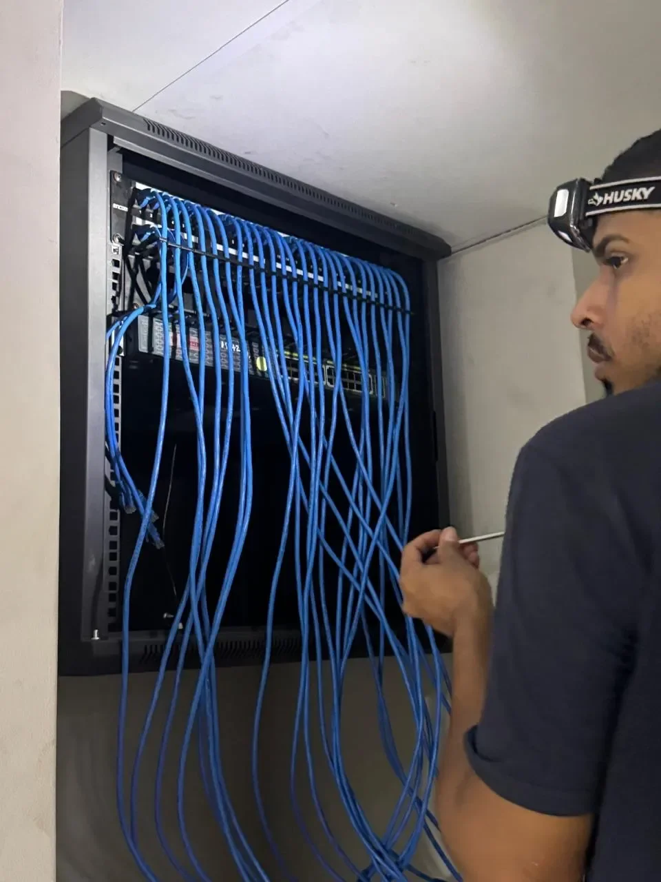 Técnico de redes trabajando en un armario de cableado, con muchas cables azul conectados a un panel de servidores o switch.