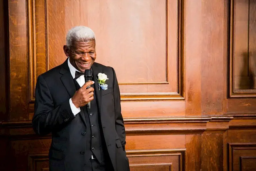 Hombre mayor con traje negro y corbata negra, sonriendo y sosteniendo un micrófono en un fondo de madera.