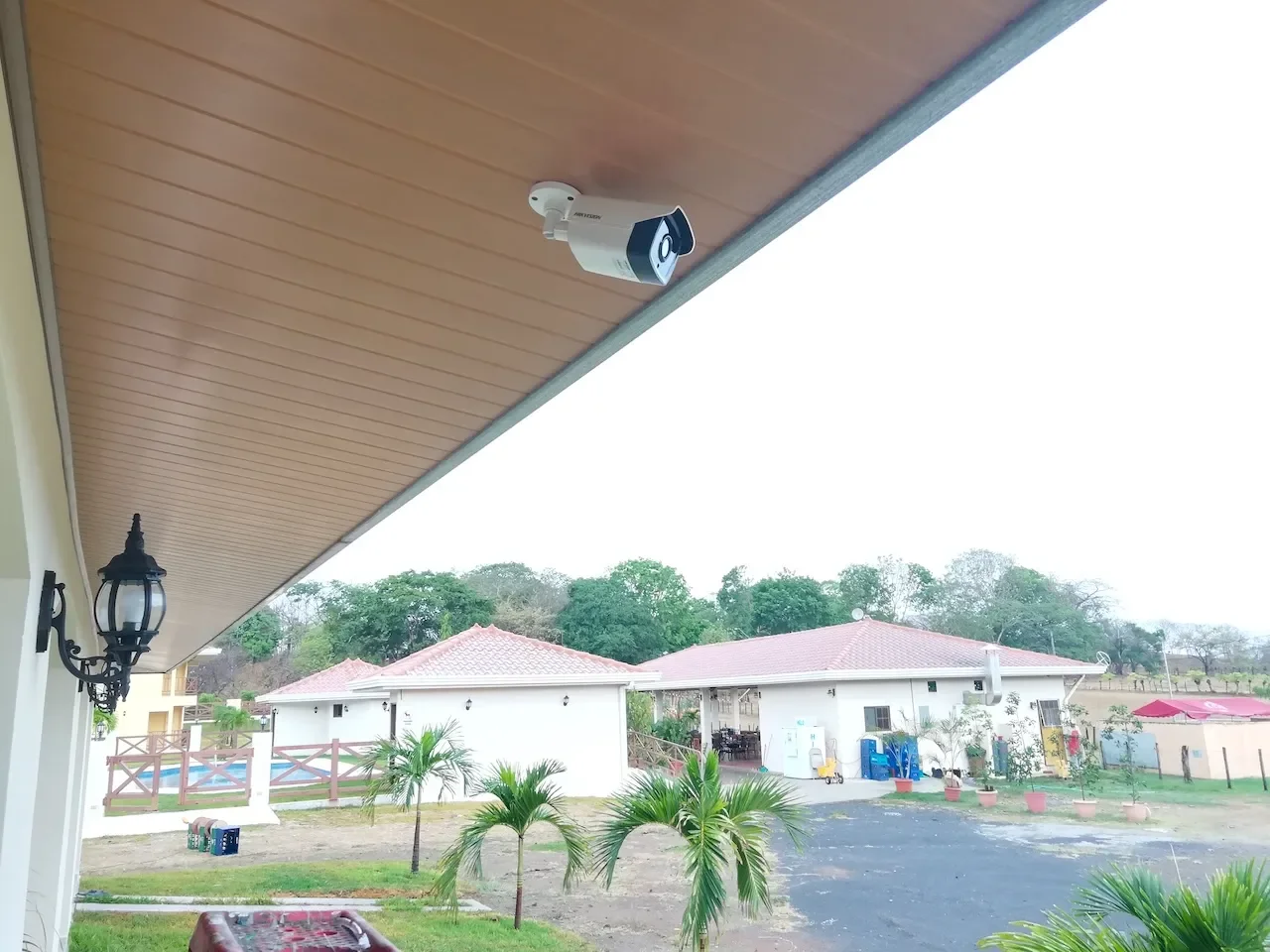 Cámara de seguridad instalada en el techo de una especie de galpón o porche con paneles de madera, vista a casas con techos rojos y áreas verdes con plantas y árboles.
