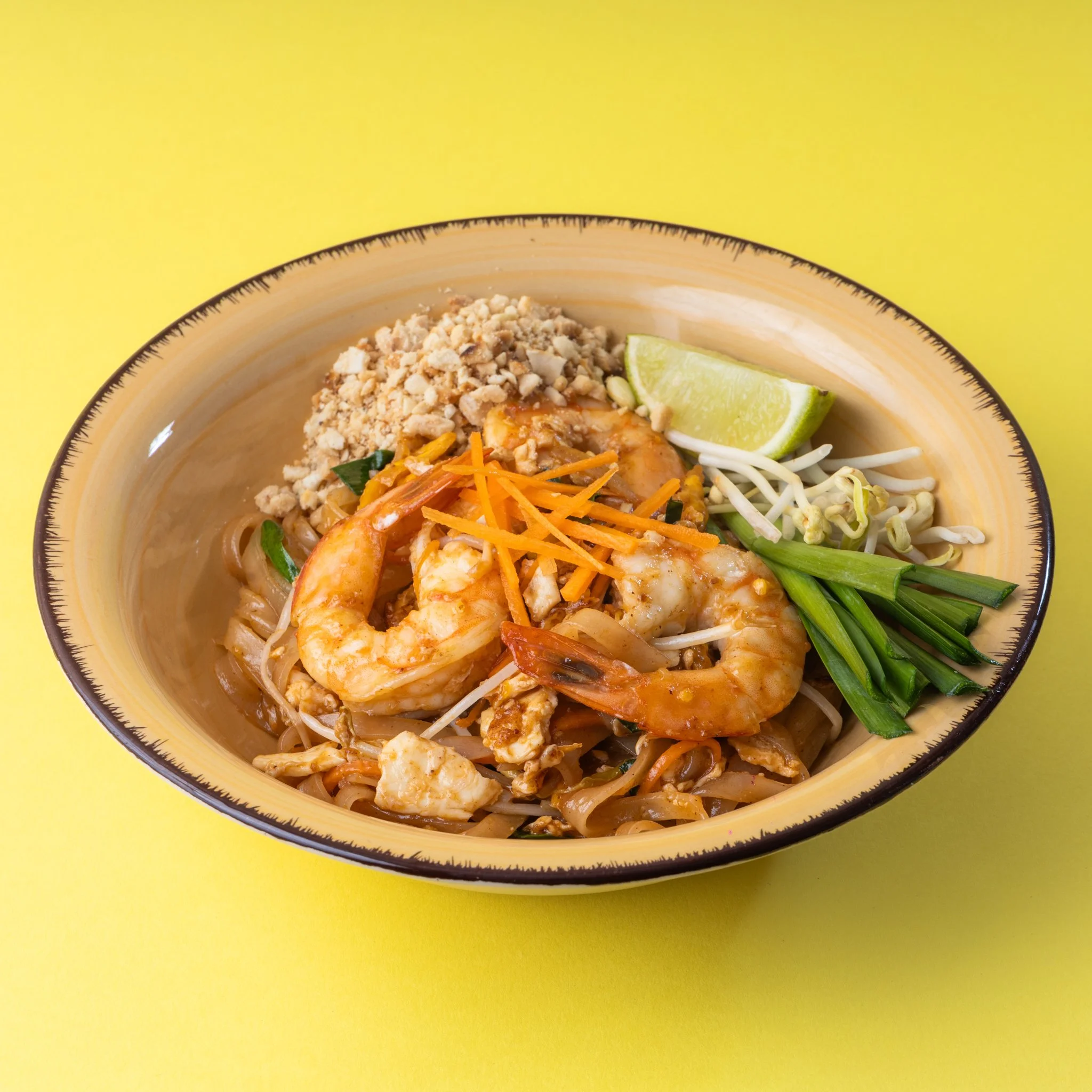 Bowl of Pad Thai with shrimp, lime wedge, bean sprouts, chopped peanuts, and shredded carrots on top, against a yellow background.