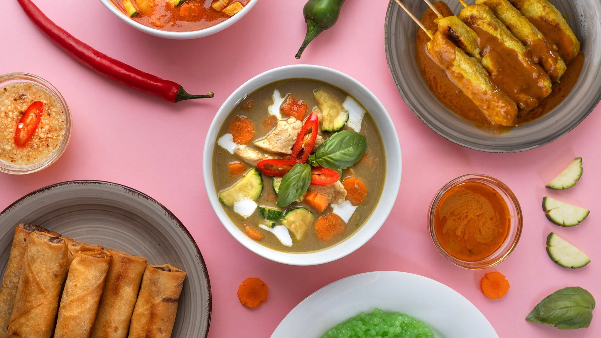 Assorted Thai dishes including spring rolls, curry, and vegetable soup on a pink surface with chili peppers, cucumber slices, and a small bowl of orange sauce.