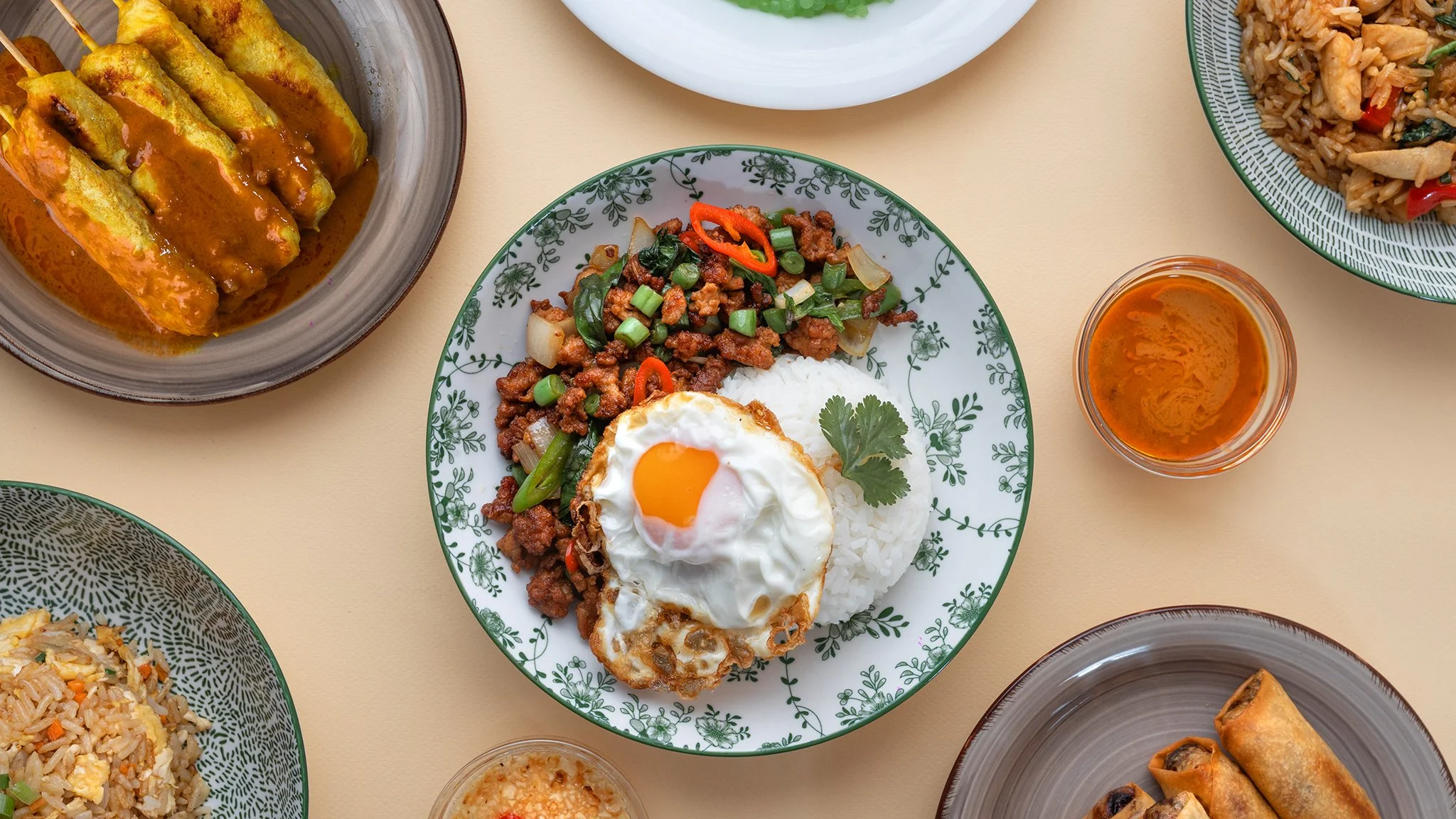 A variety of Asian dishes on a light-colored table, including fried rice, spring rolls, satay skewers, and a plate with rice, a fried egg, and ground meat stir-fry, with small bowls of sauce