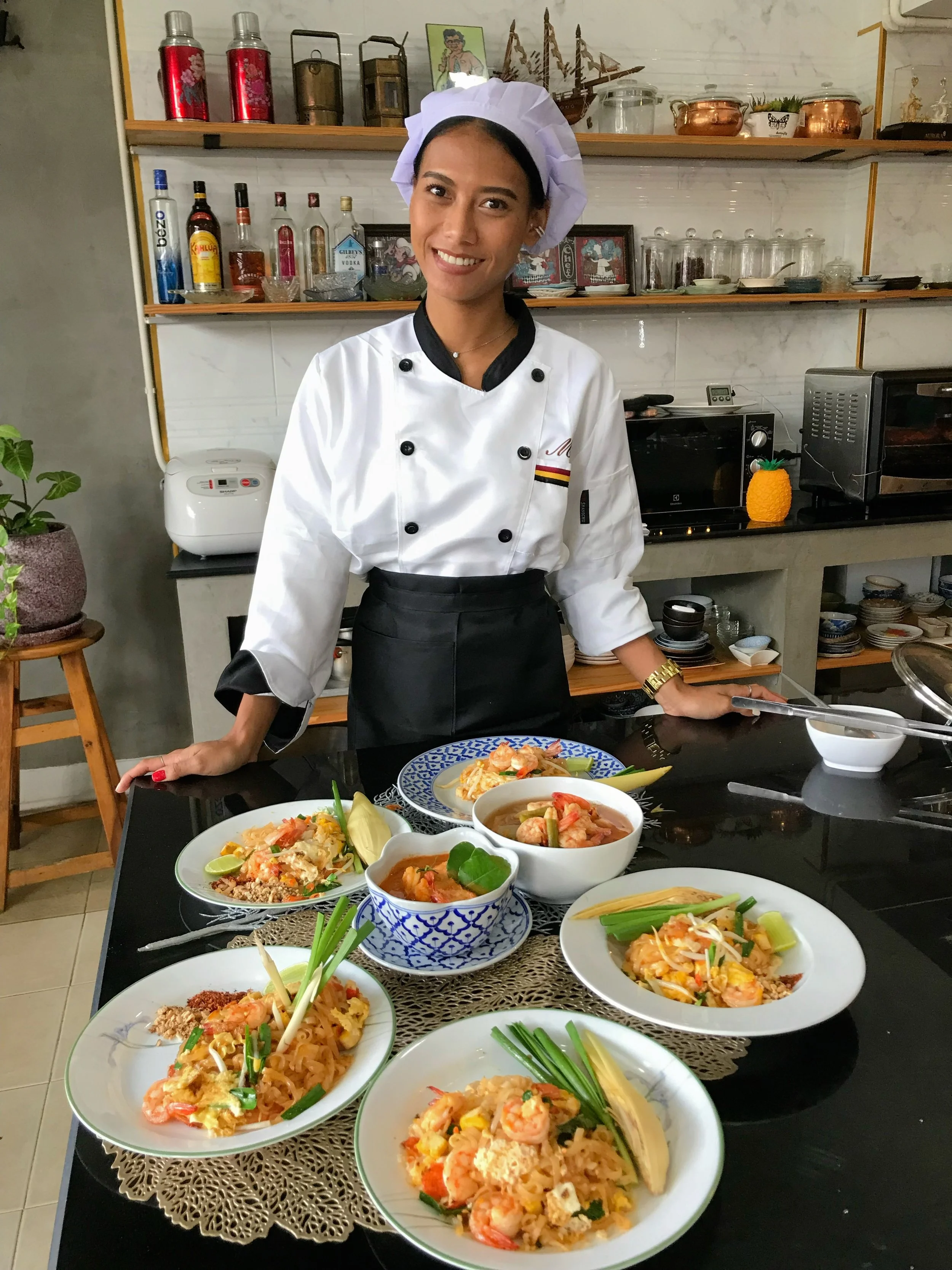 A woman dressed in a chef's uniform and a white chef's hat stands behind a table with various plates of colorful Thai dishes, including shrimp and noodles, in a kitchen with shelves of bottles, bowls, and kitchen appliances.
