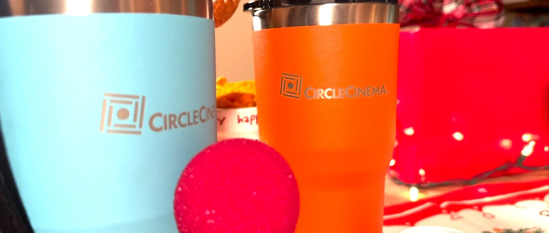 Close-up of two Circle Cinema-branded tumblers, one in blue and the other in orange. The blue one has a red Rudolph nose. There are also blurred background objects with Christmas lights and decorations.