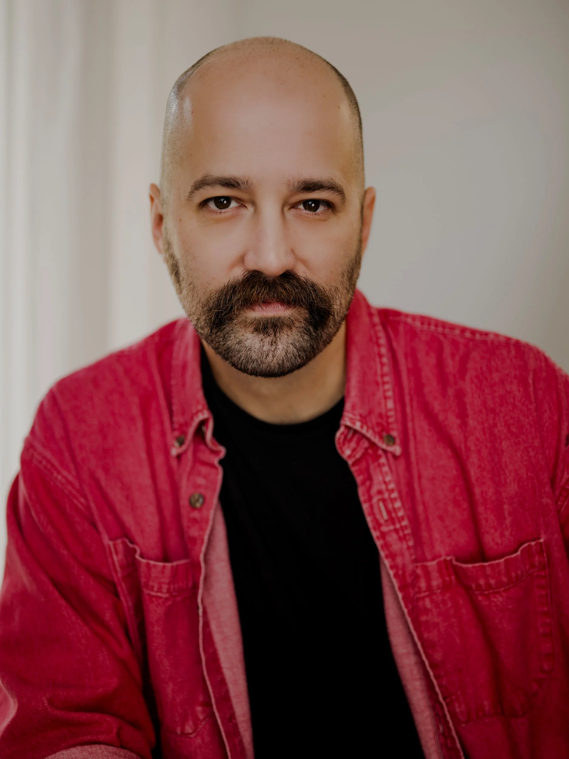 Portrait of Tyler Huffman, a man with a bald head, stubble, and a mustache, wearing a red shirt over a black T-shirt, looking directly at the camera against a plain, neutral background.