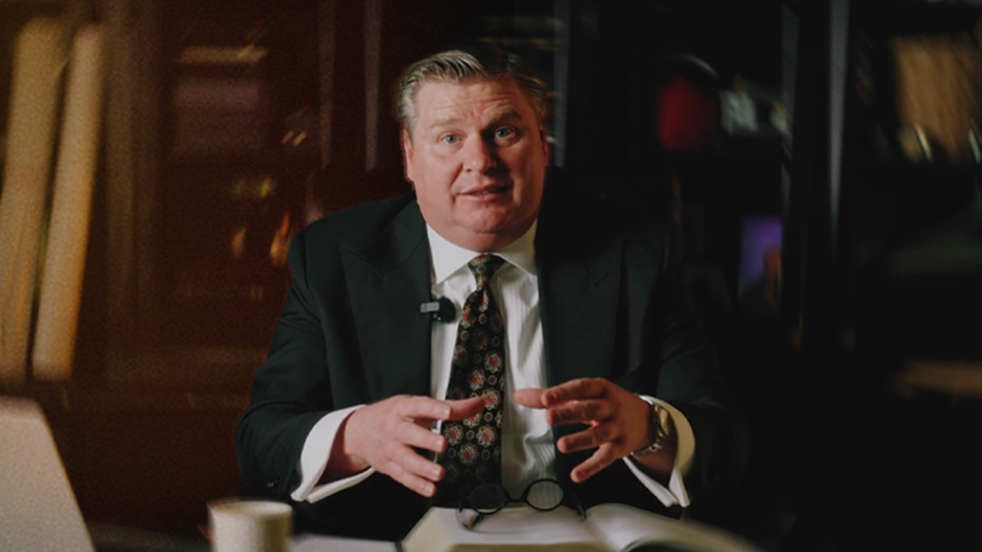 A man in a black suit and patterned tie sitting at a desk, gesturing with his hands, in a dimly lit office or library setting.