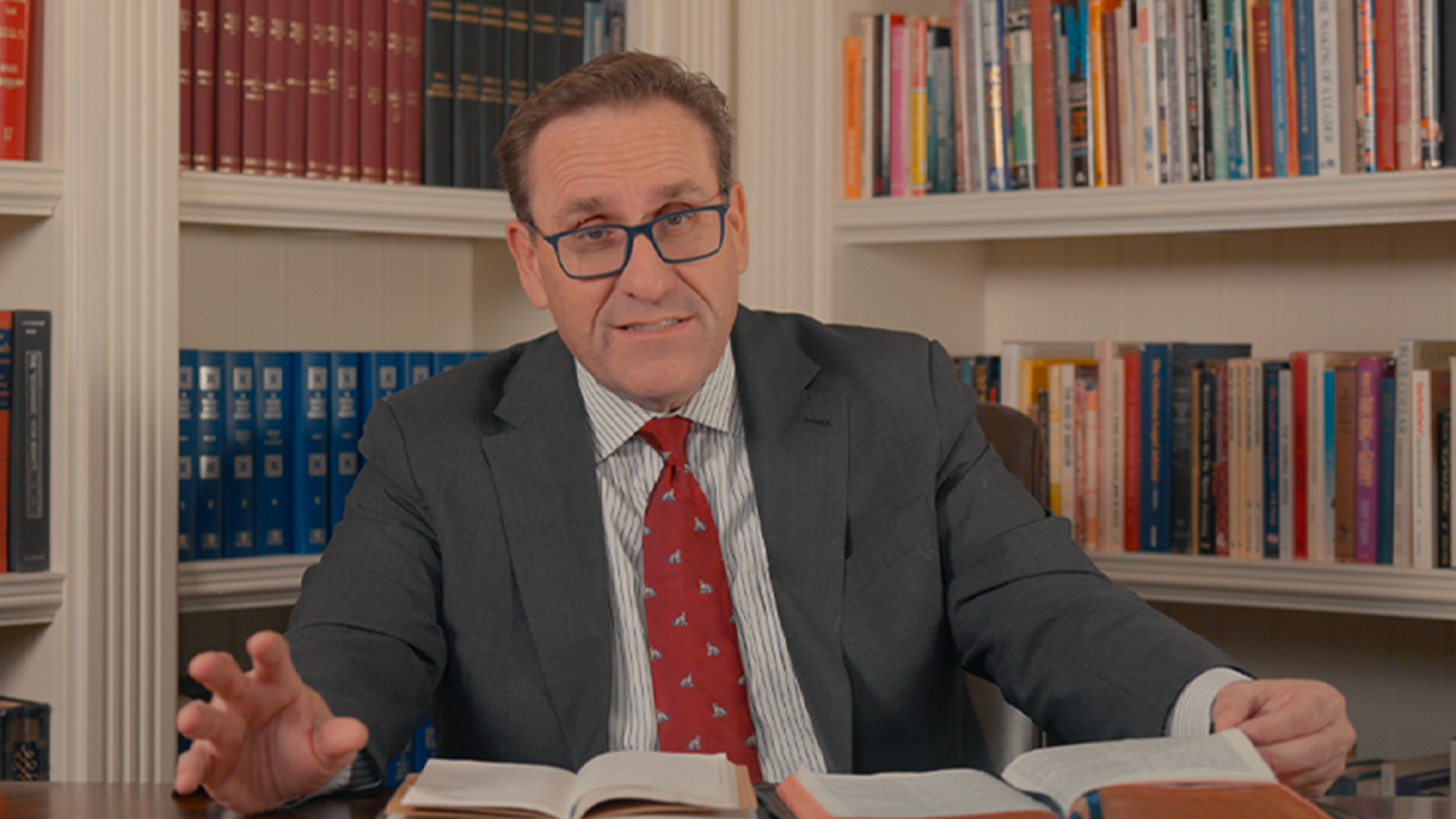 A man in a gray suit and red tie sitting at a table with an open book, gesturing with his left hand, in front of a bookshelf filled with books.