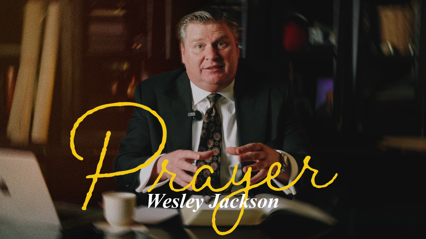 A man in a suit and tie sitting at a desk with a book, coffee cup, and laptop, gesturing with his hands during a video interview.