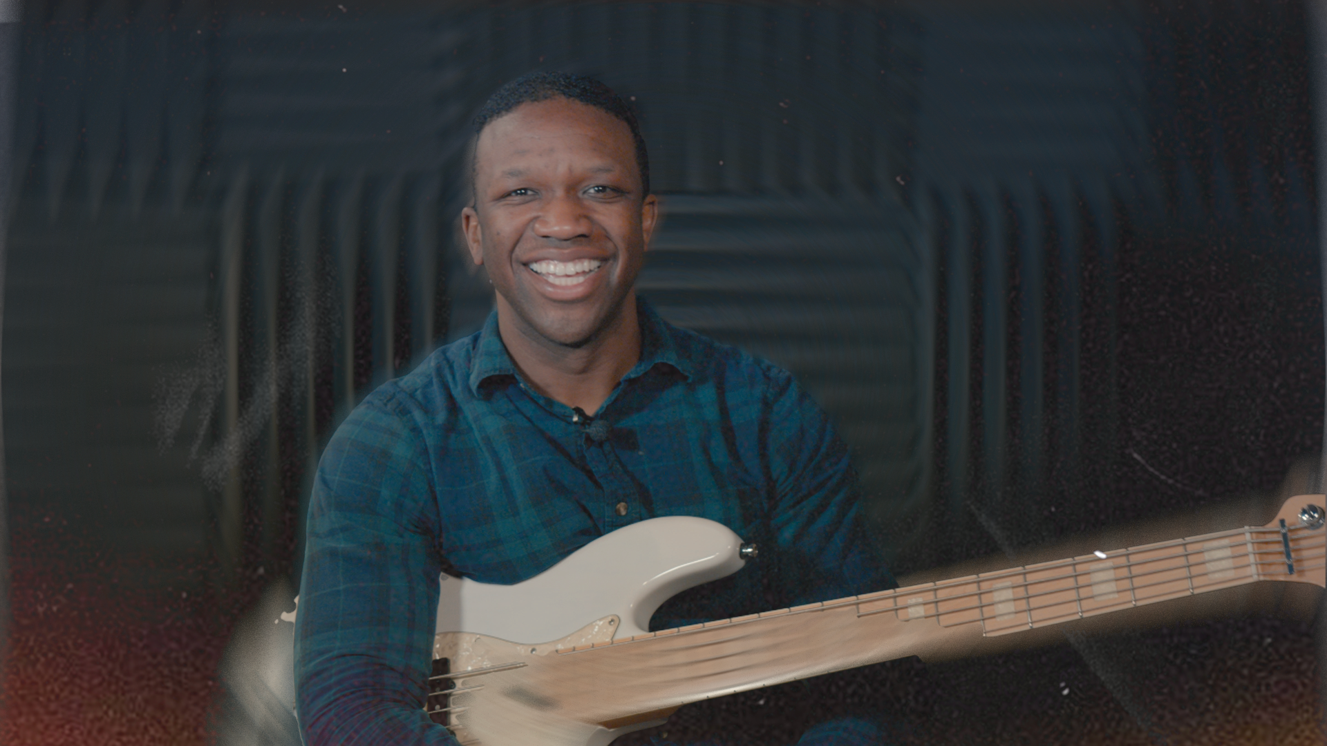 A smiling man with short dark hair wearing a blue plaid shirt, holding a white electric bass guitar in a music studio with soundproofing panels in the background.