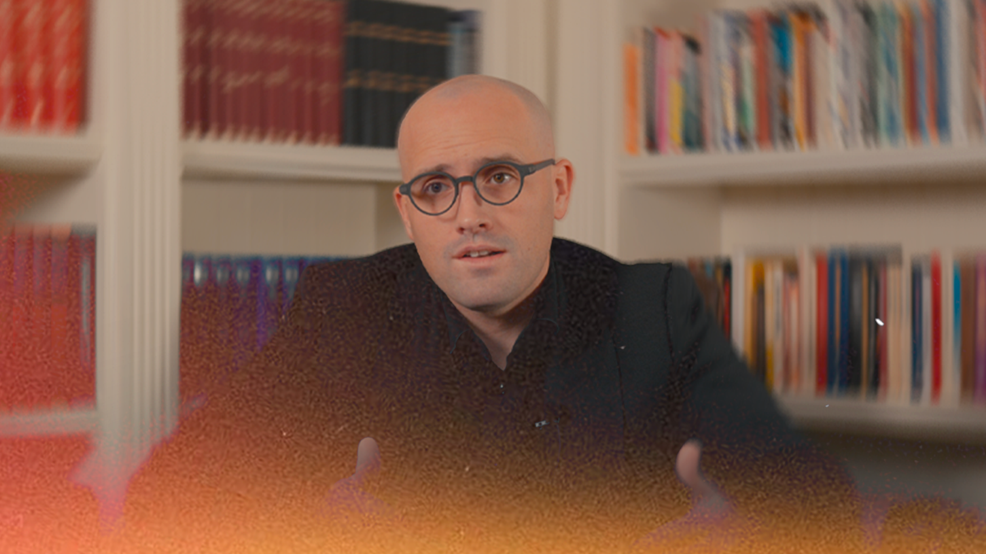 A man with a bald head and glasses sitting in front of a bookshelf filled with books, wearing a dark shirt, speaking or explaining, in an indoor setting.