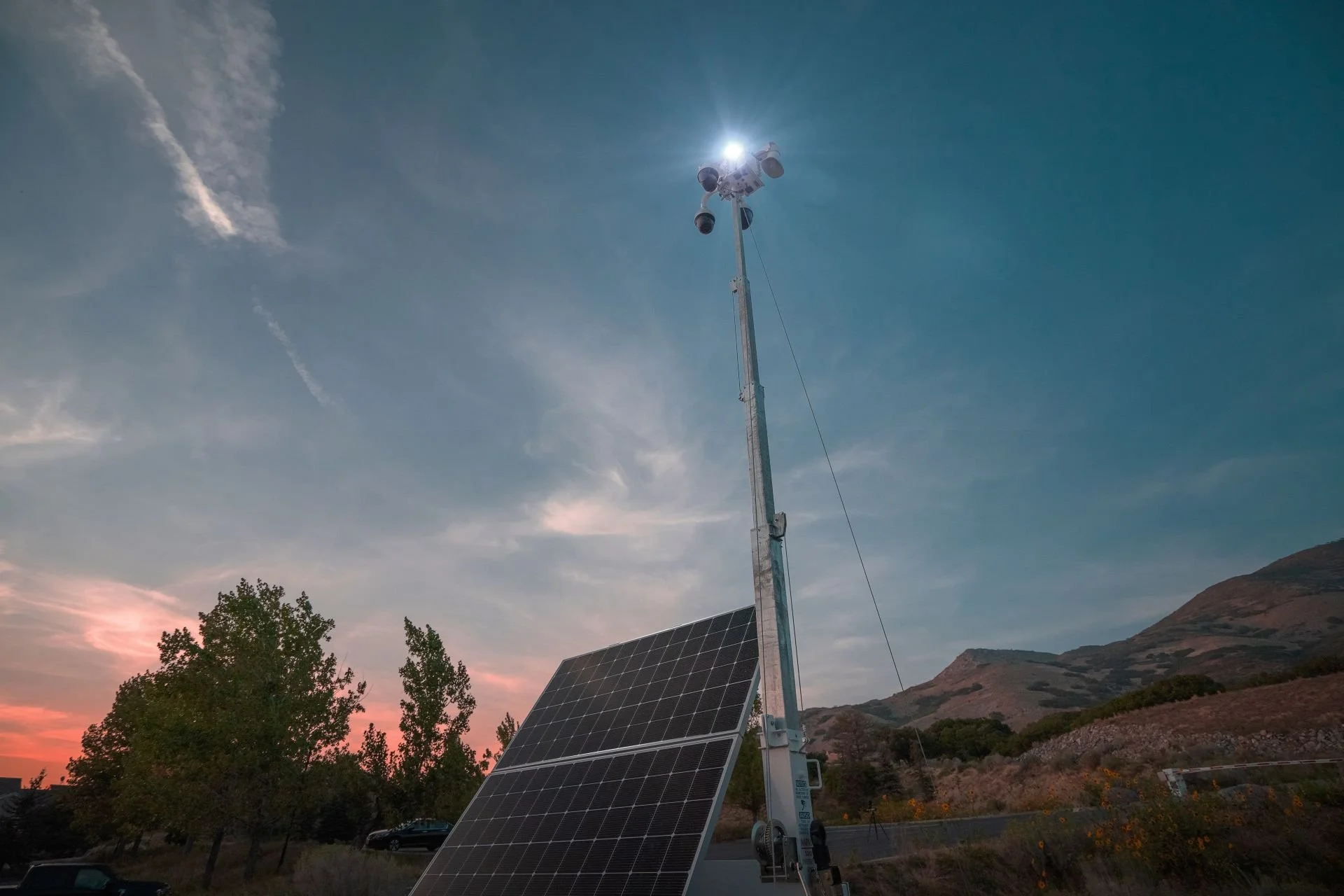 A solar panel and a tall tower with cameras and sensors under a partly cloudy sky during sunset.