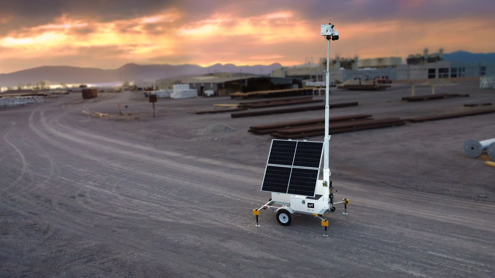 A mobile solar-powered surveillance camera system with solar panels and a backup generator on a dirt road during sunset, with a construction site and mountains in the background.