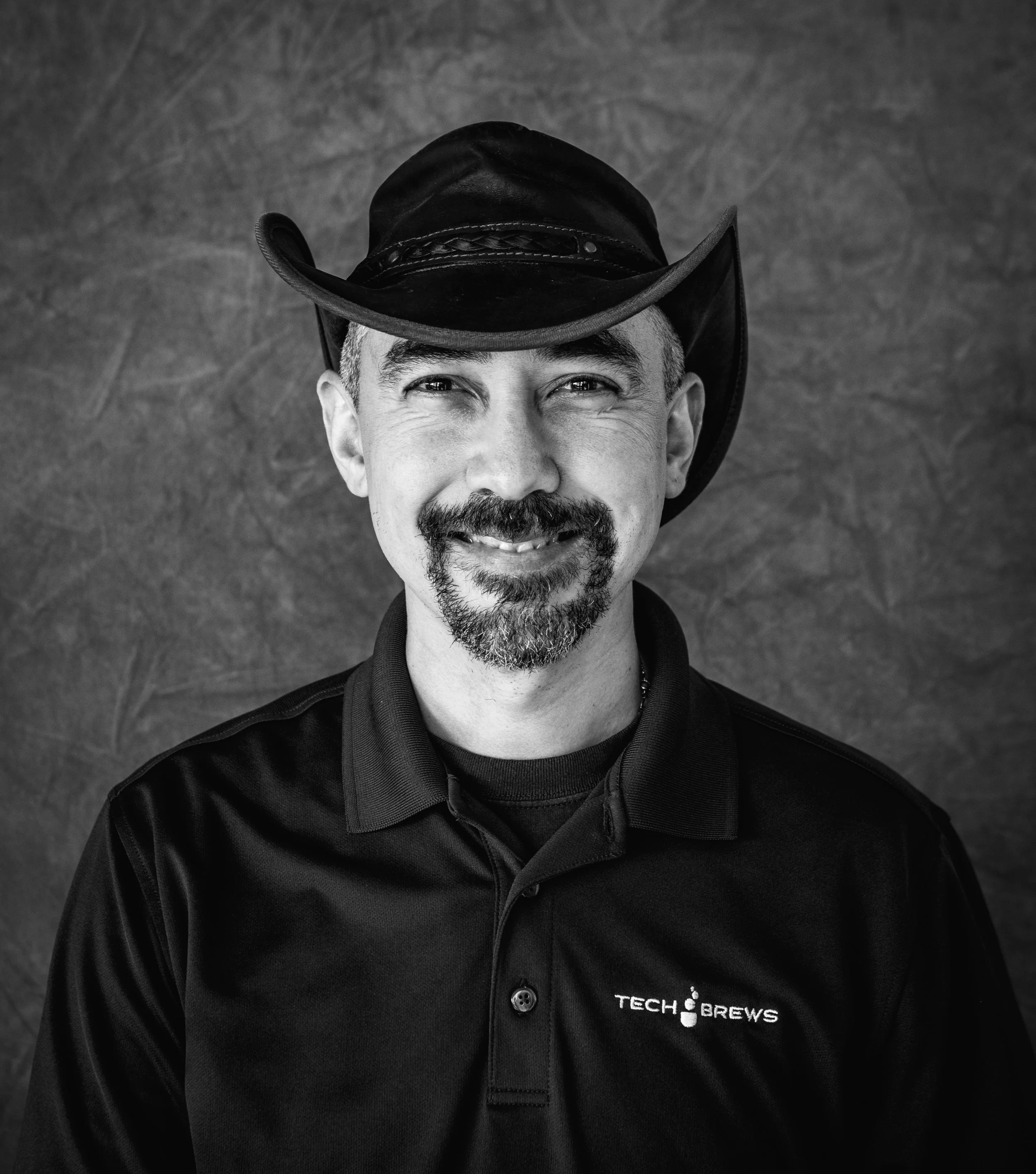 Black and white portrait of a man wearing a cowboy hat and a black polo shirt with the 'Tech Brews' logo, smiling and looking at the camera.