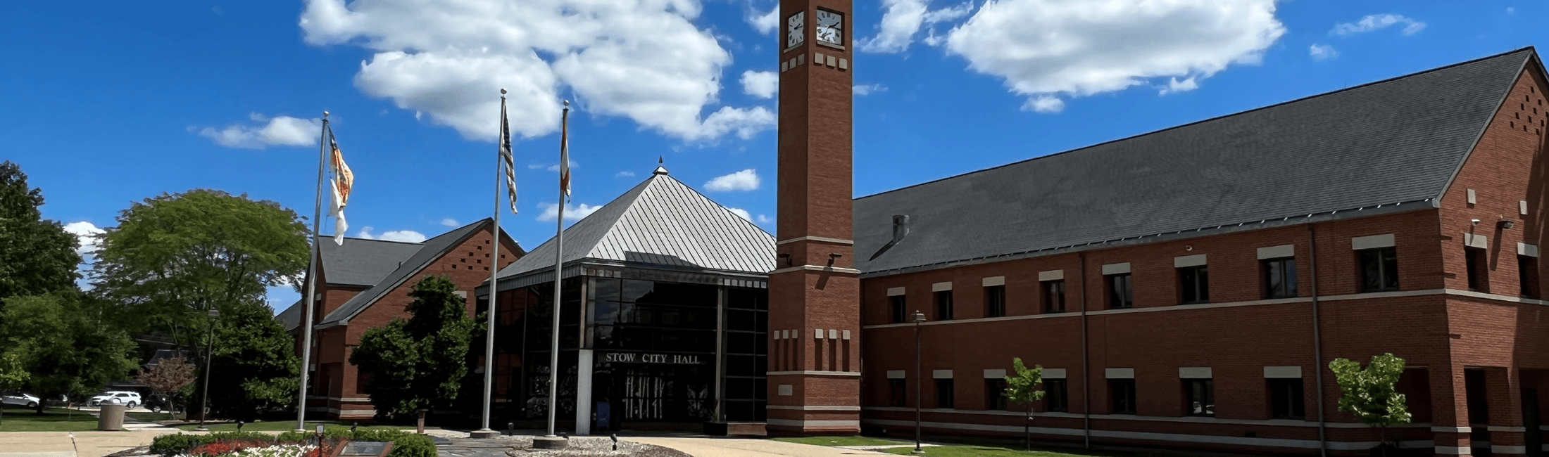 A large brick city hall building with a clock tower, three flagpoles with flags, a glass entrance, surrounded by trees and a clear blue sky with clouds.