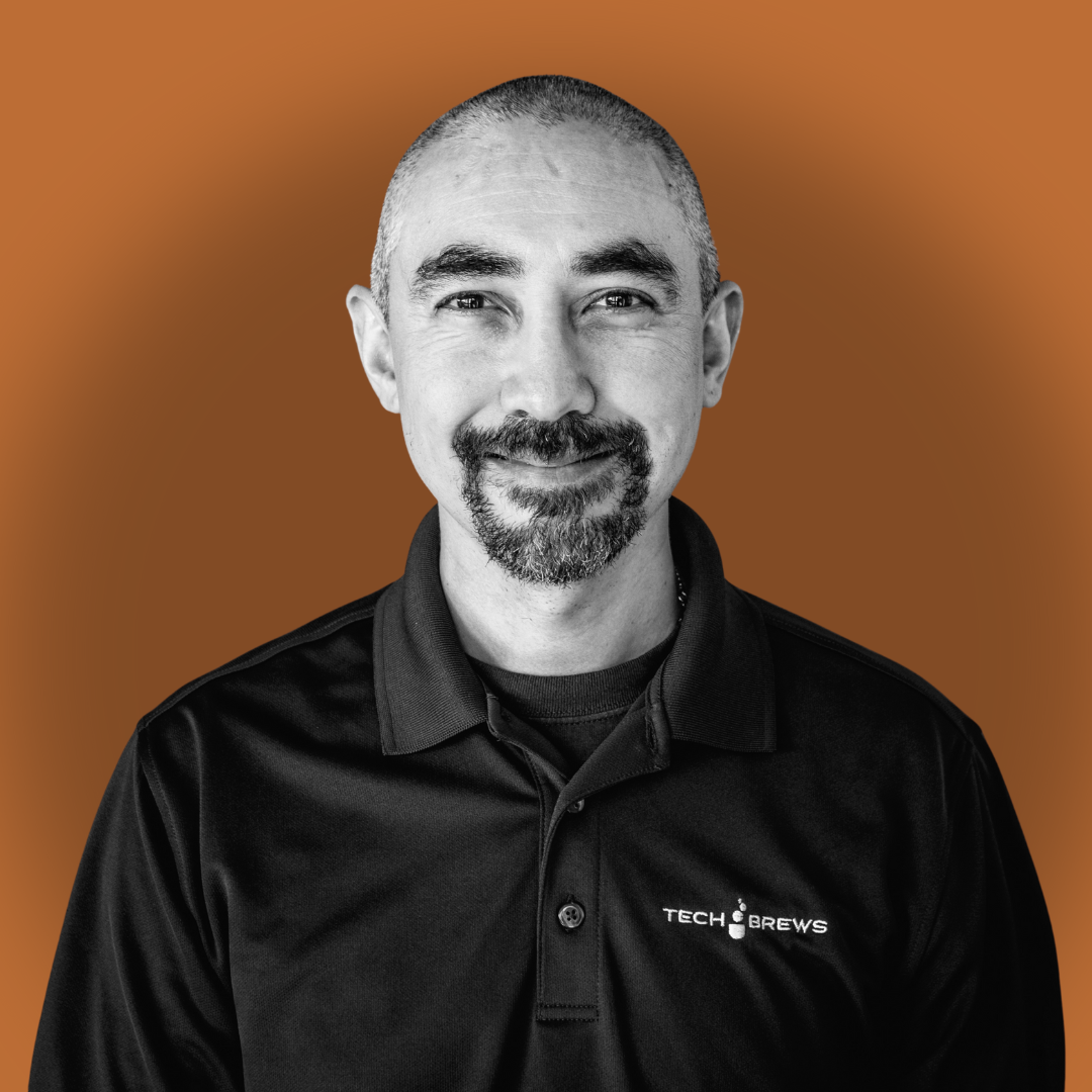 A man with short hair and a goatee, smiling, wearing a black collared shirt with a 'Tech Brews' logo on it, over a brown background.
