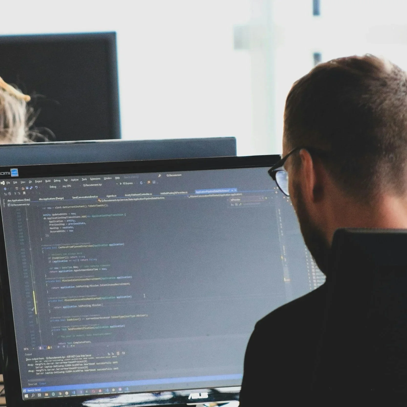 A man with glasses working on a computer with code on the screen. Behind him is a bright window, and part of another person’s head is visible on the left.