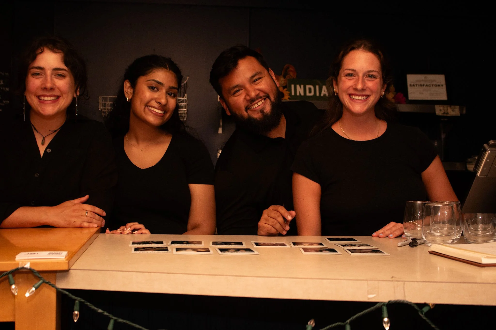 Four people smiling behind a table with photographs, glassware, and a laptop in an intimately lit indoor setting.