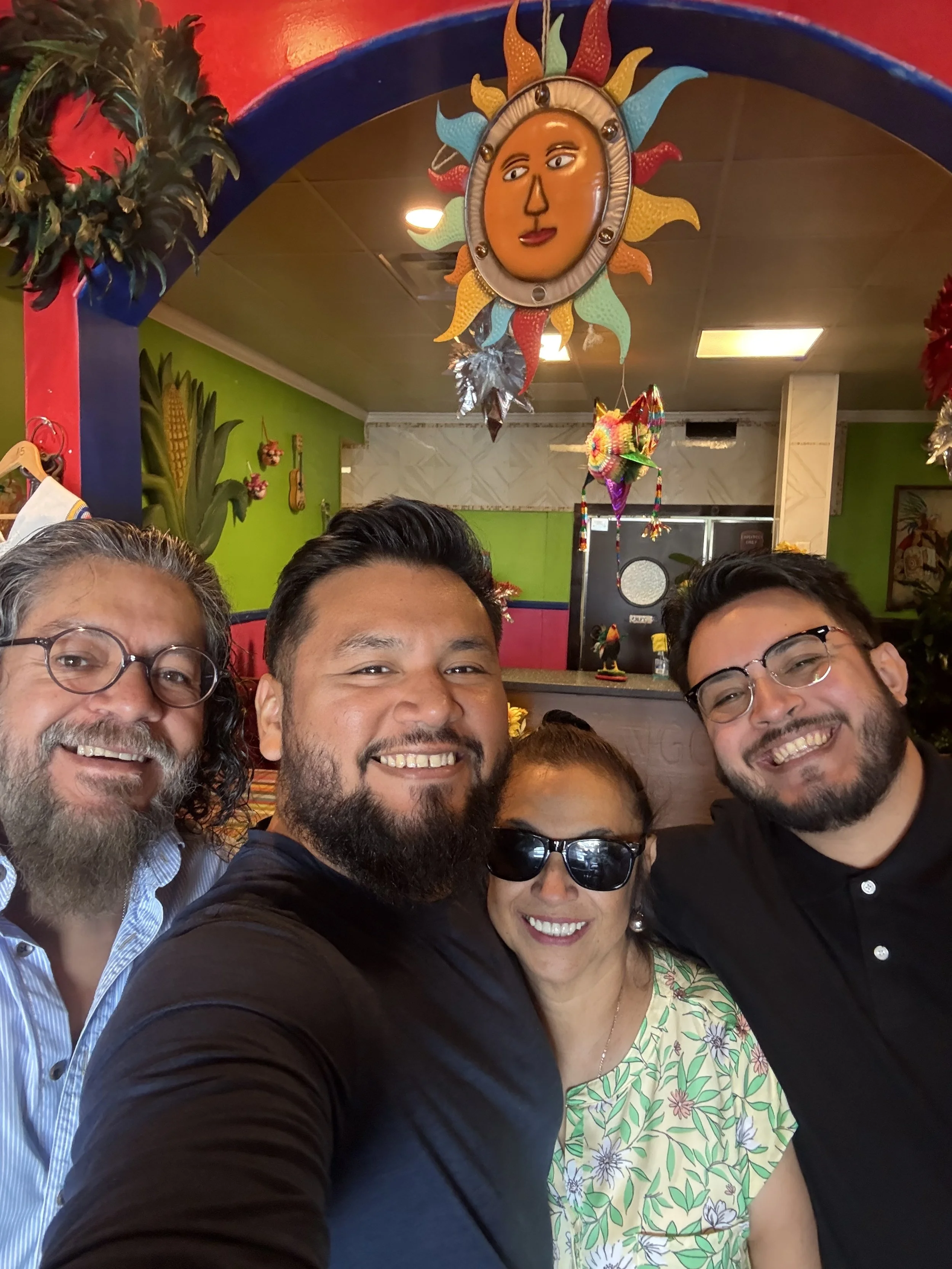Four people smiling for a selfie in a colorful restaurant decorated with a sun face and vibrant decorations.
