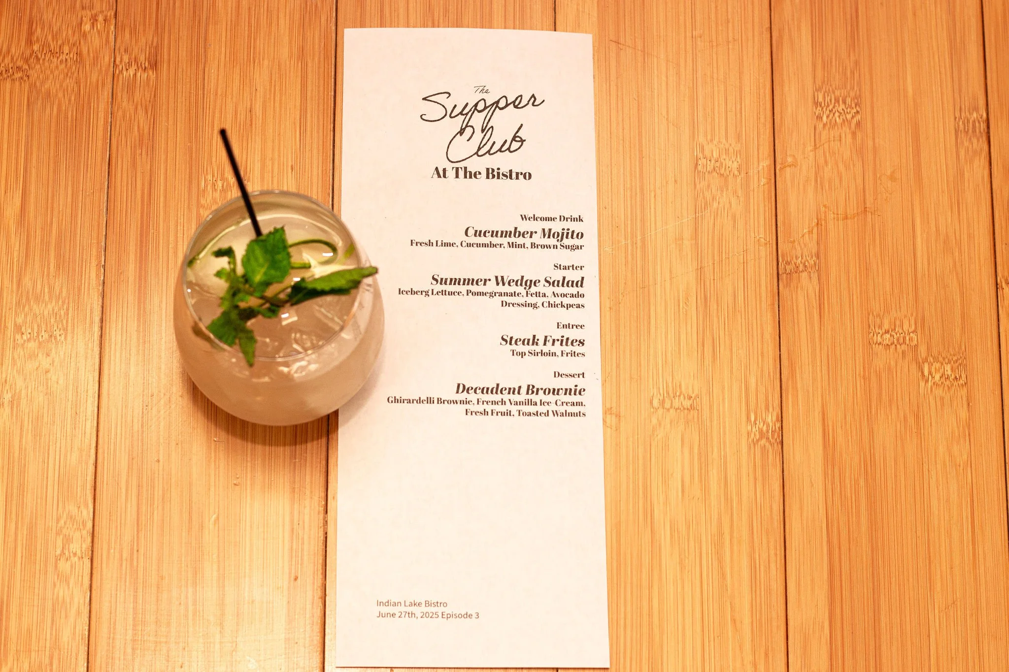 A cocktail drink garnished with mint leaves, served with a straw, placed on a wooden table next to a printed menu for a supper club at the bistro