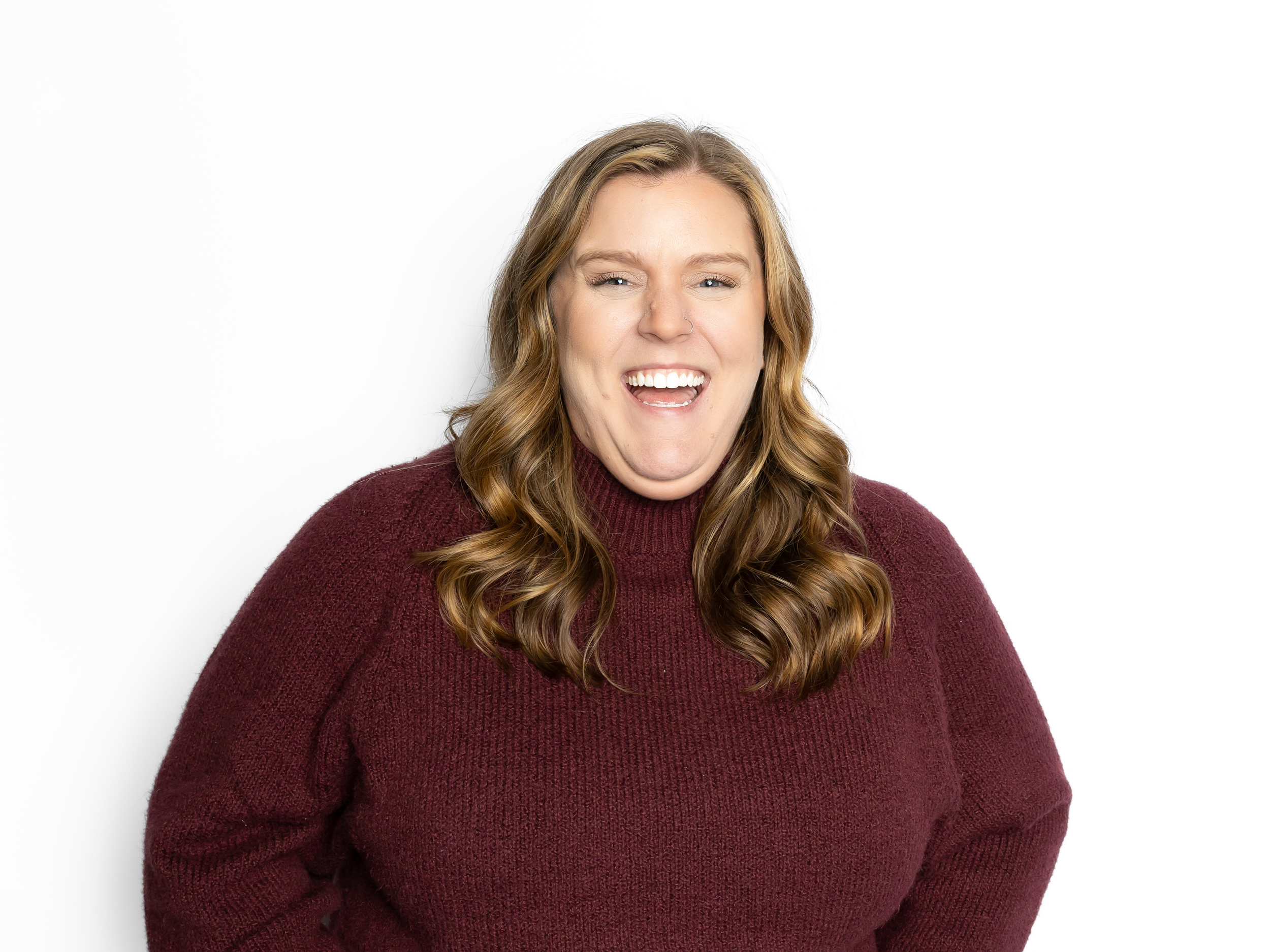 A woman with long wavy brown hair wearing a maroon sweater, smiling with one eye closed and showing teeth, against a plain white background.