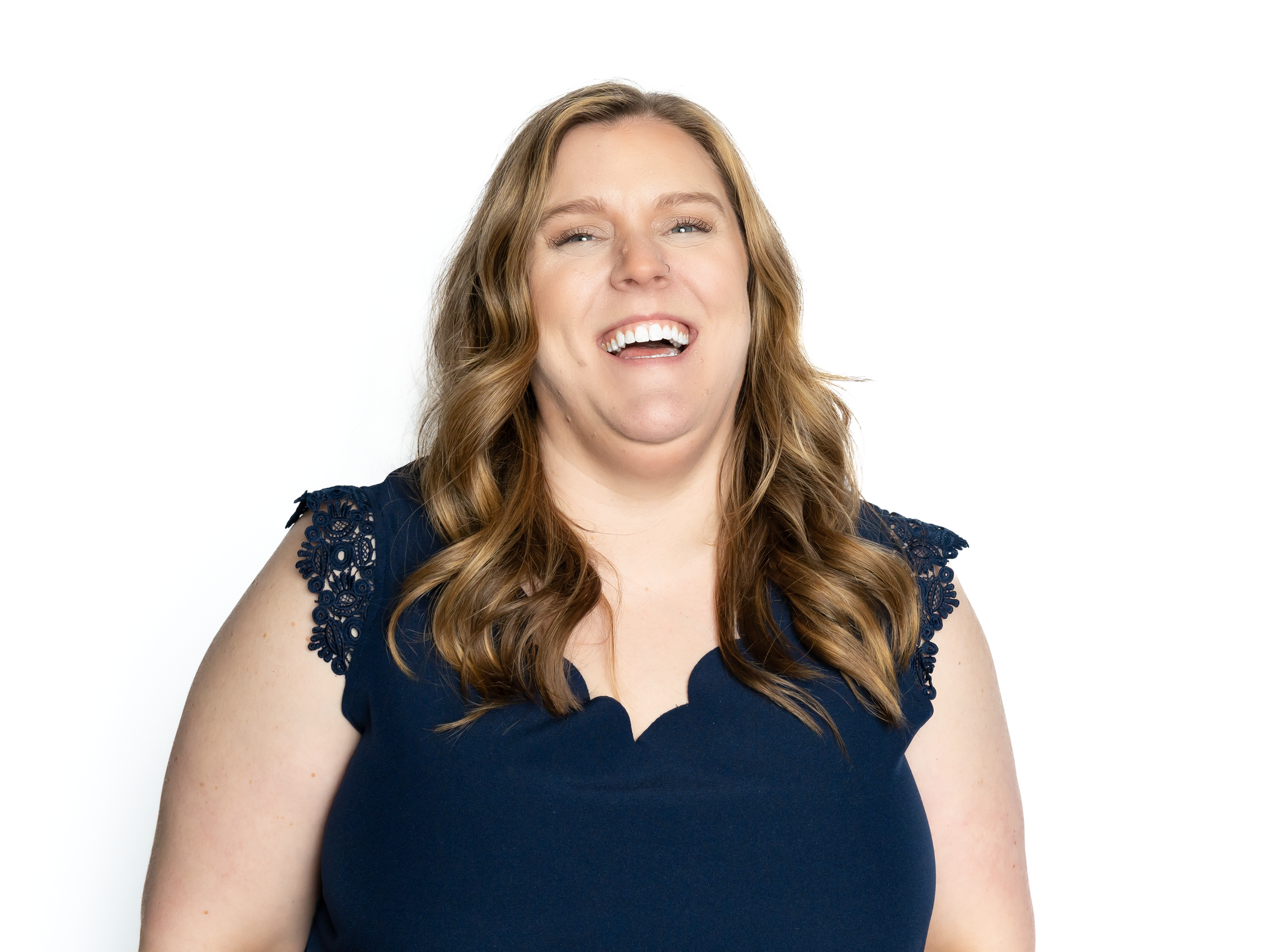 A woman with light brown hair wearing a navy blue top with lace details on the shoulders, smiling and laughing.