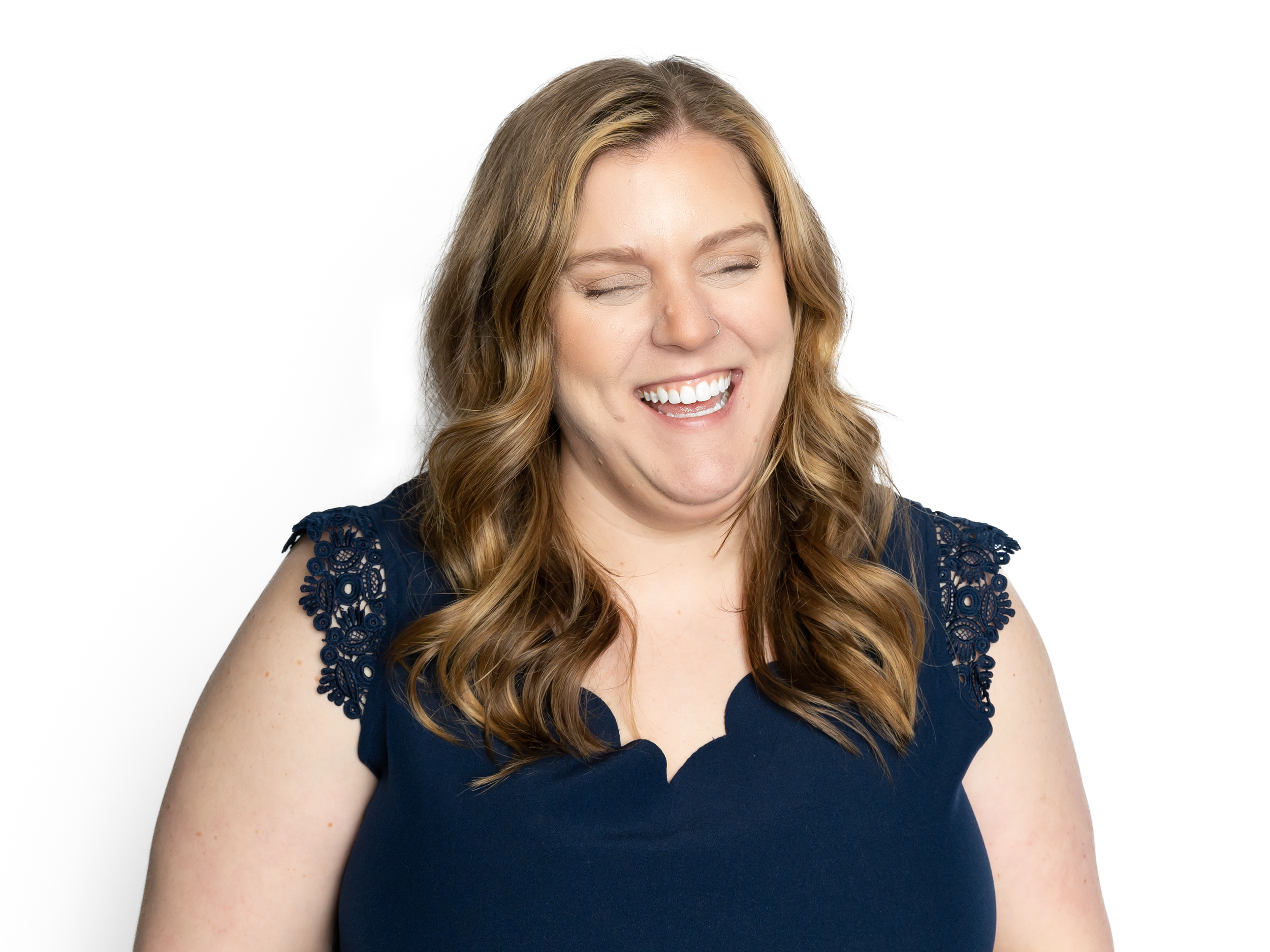 A woman with long wavy brown hair wearing a navy blue top with lace shoulder details, smiling with her eyes closed.